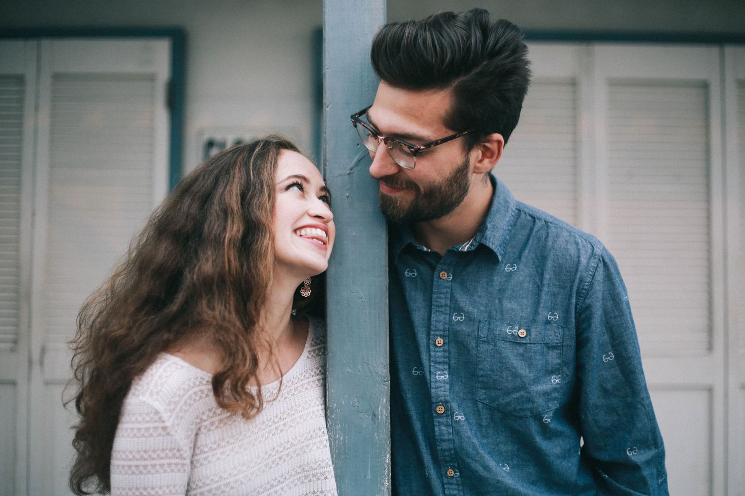 Couple smiles at each other leaning against a pillar outdoors in casual attire.