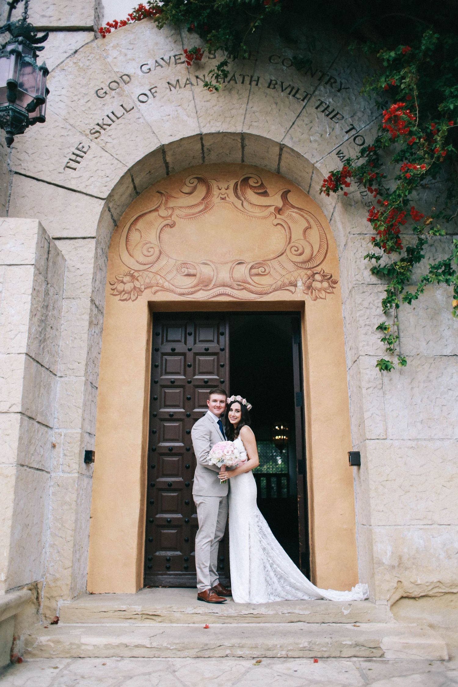 Bride and groom posing together at arched stone doorway with decorative scrollwork and red flowers.