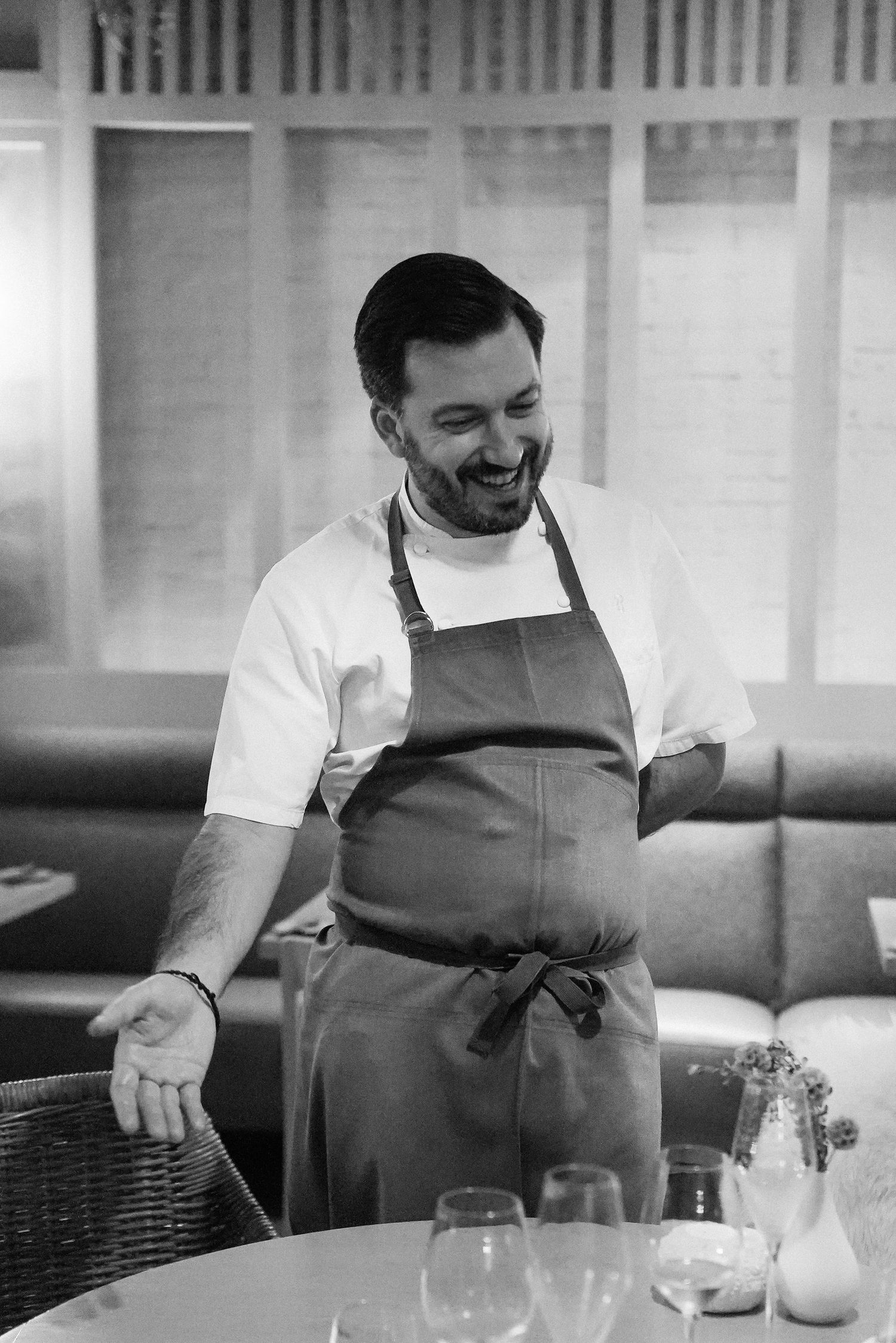 Smiling chef in white shirt and brown apron standing in elegant restaurant dining room.