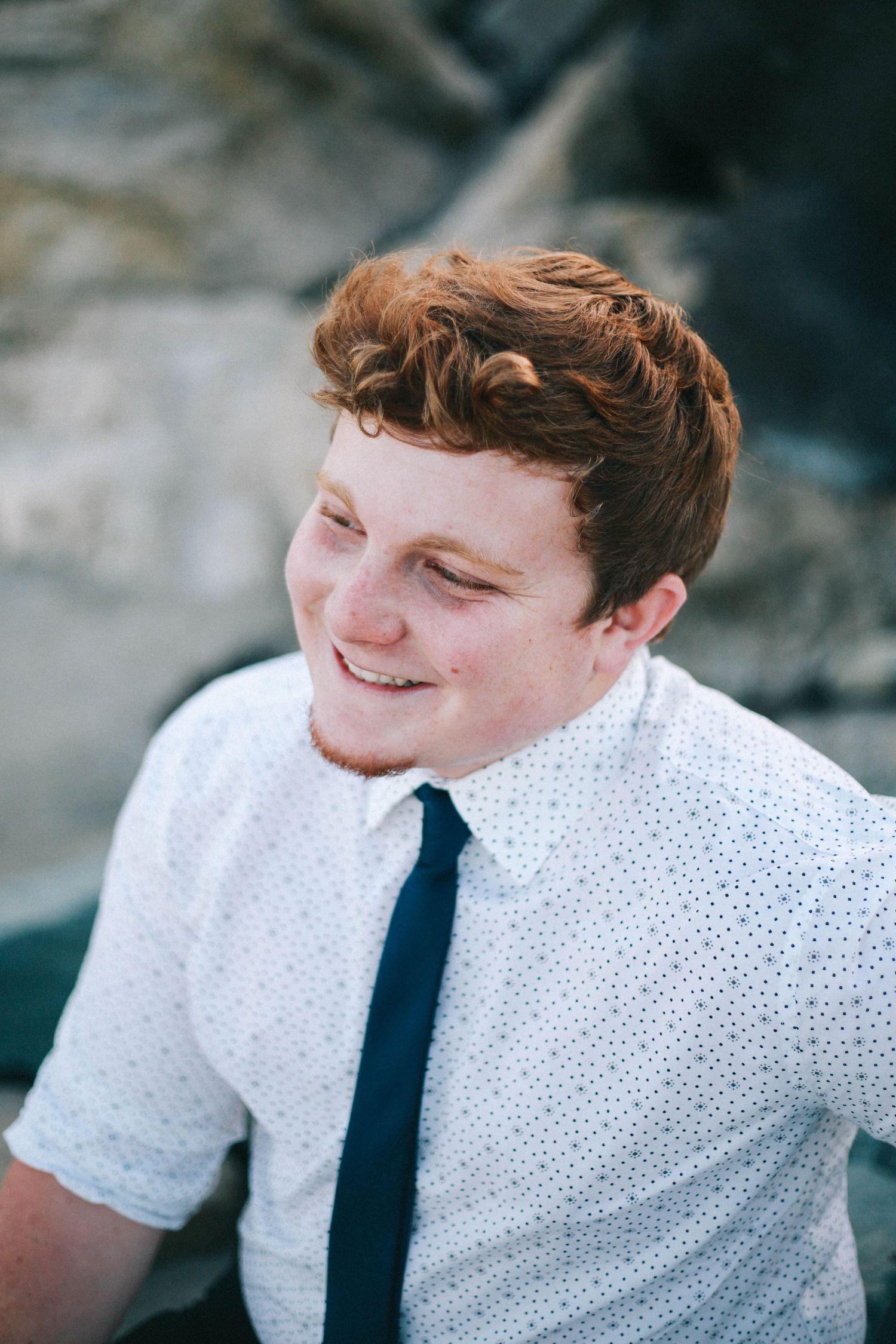 Smiling redhead young man in white shirt and navy tie sits outdoors in natural light.