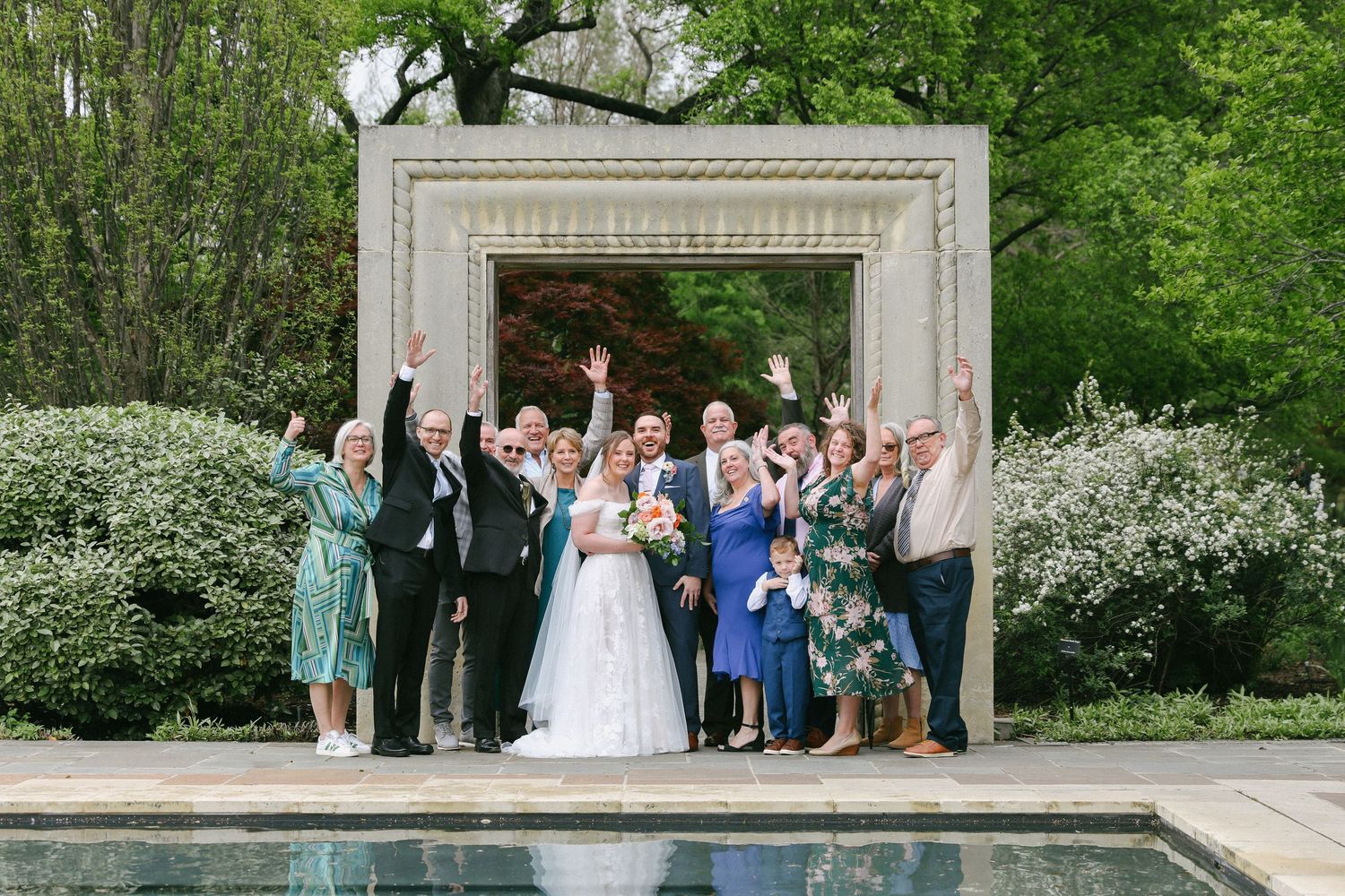 Wedding party celebrating outdoors under stone arch near pool surrounded by spring blooms.