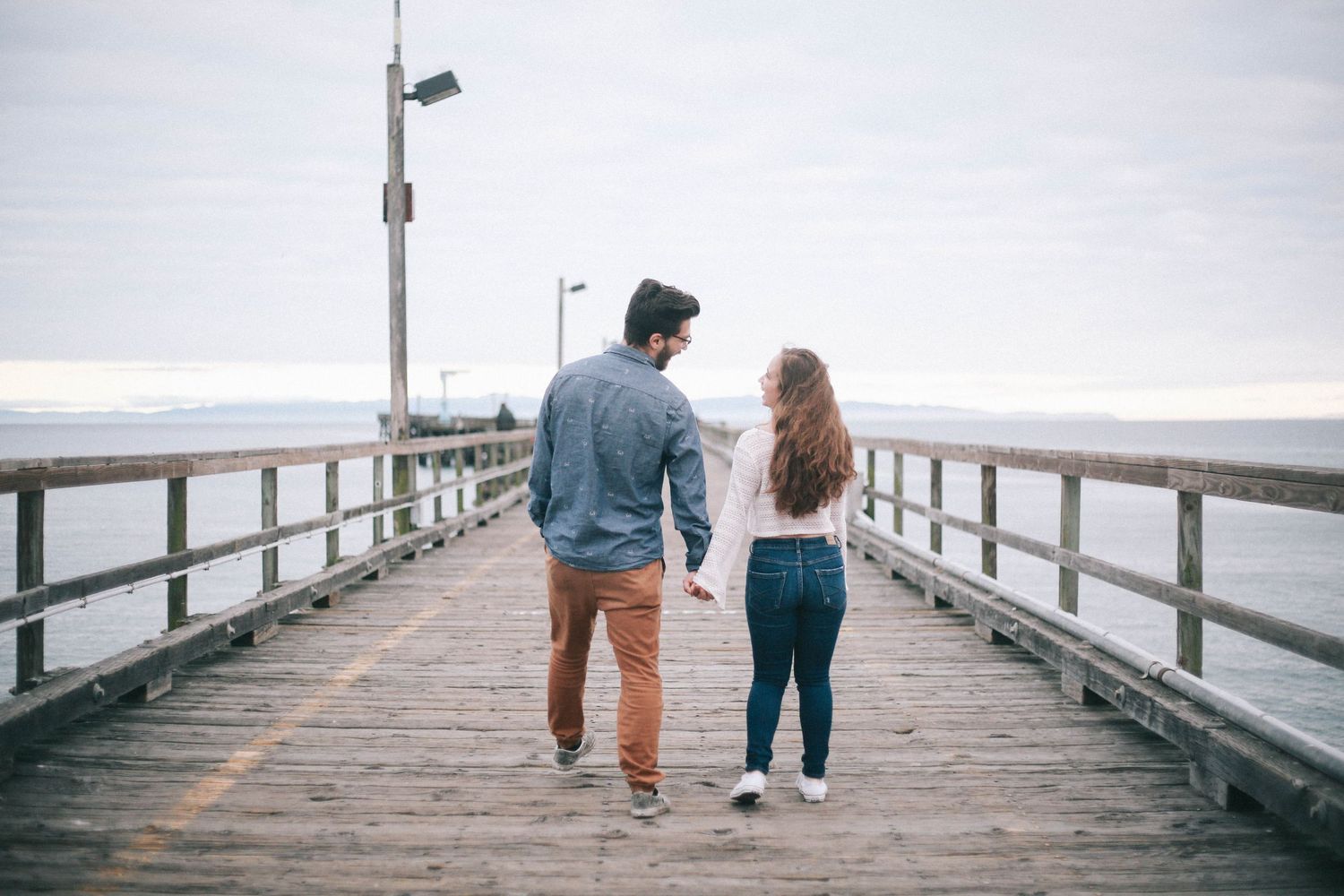 Couple holding hands walking on a wooden pier overlooking the ocean on a cloudy day.
