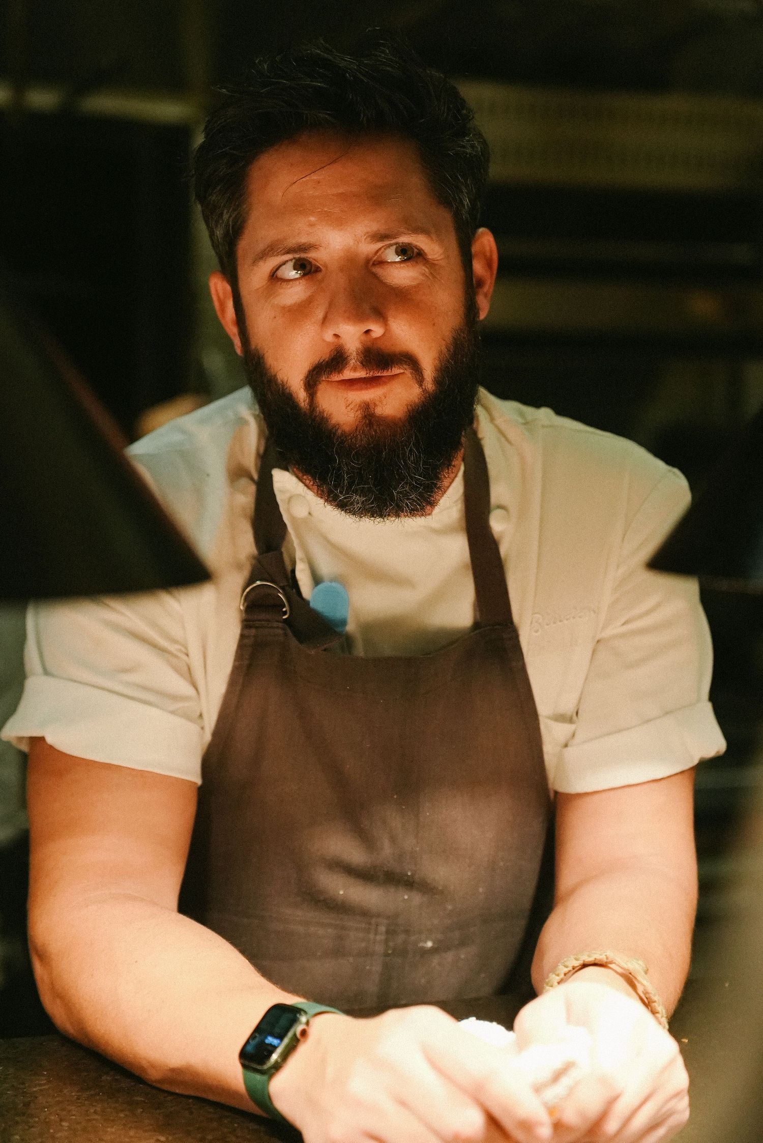 Bearded chef in brown apron leaning on counter in professional kitchen setting.