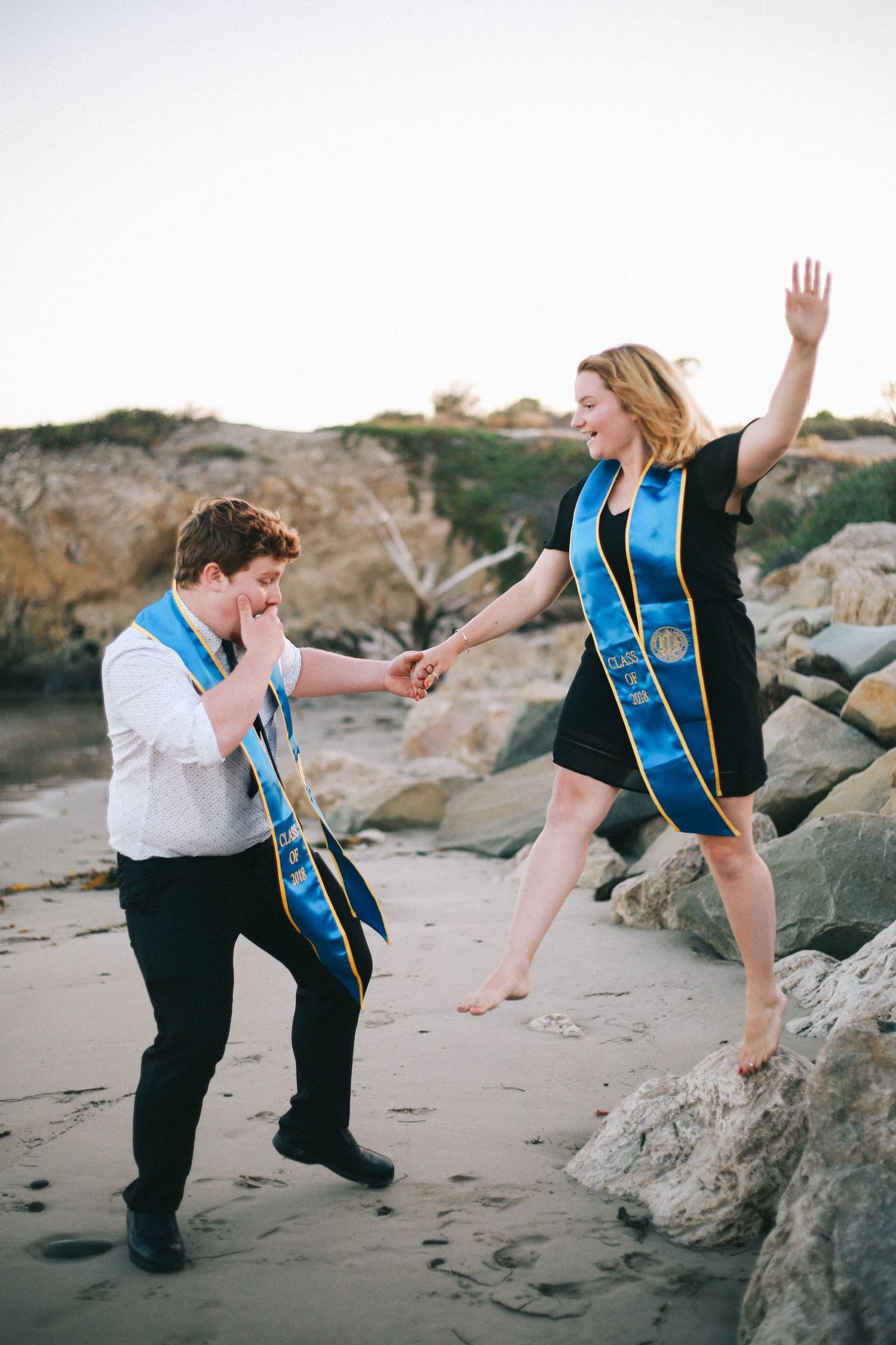 Two UCSB graduates in blue stoles celebrating on a rocky beach, one jumping joyfully.