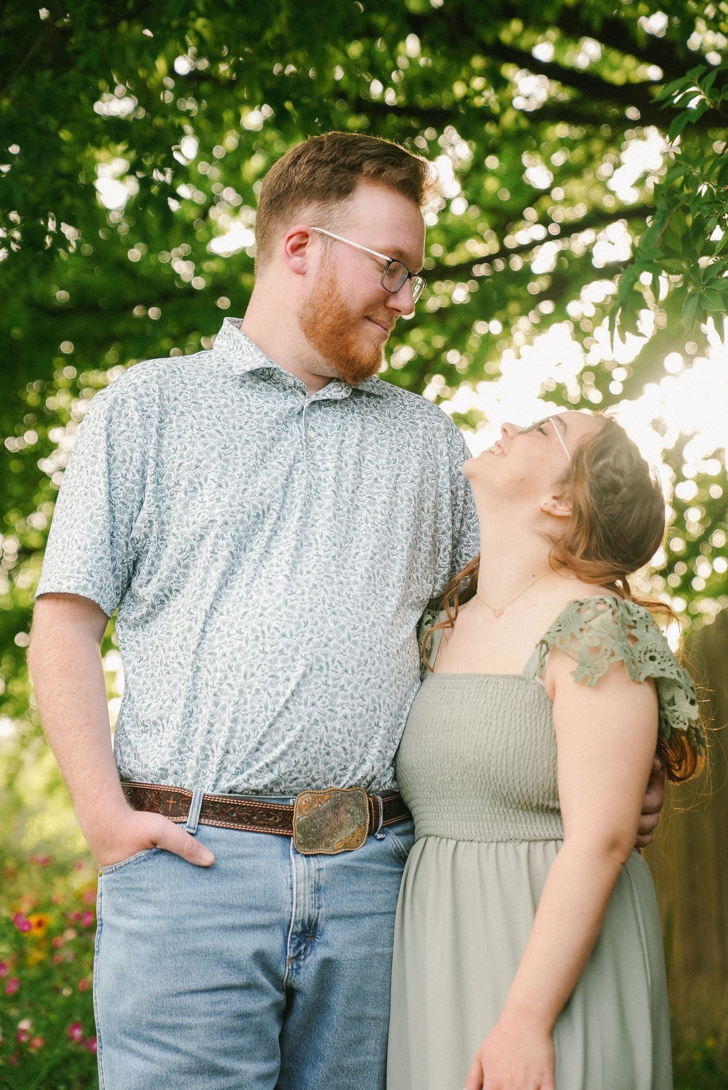 Engaged couple smiling at each other under lush green trees in golden hour sunlight.