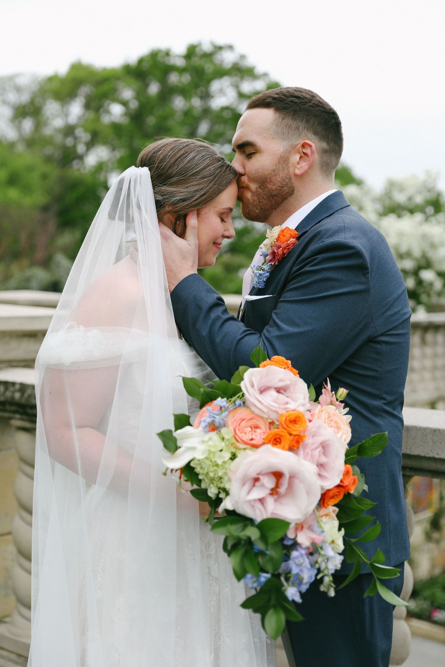 Groom kissing bride on forehead while she holds colorful spring wedding bouquet outdoors.