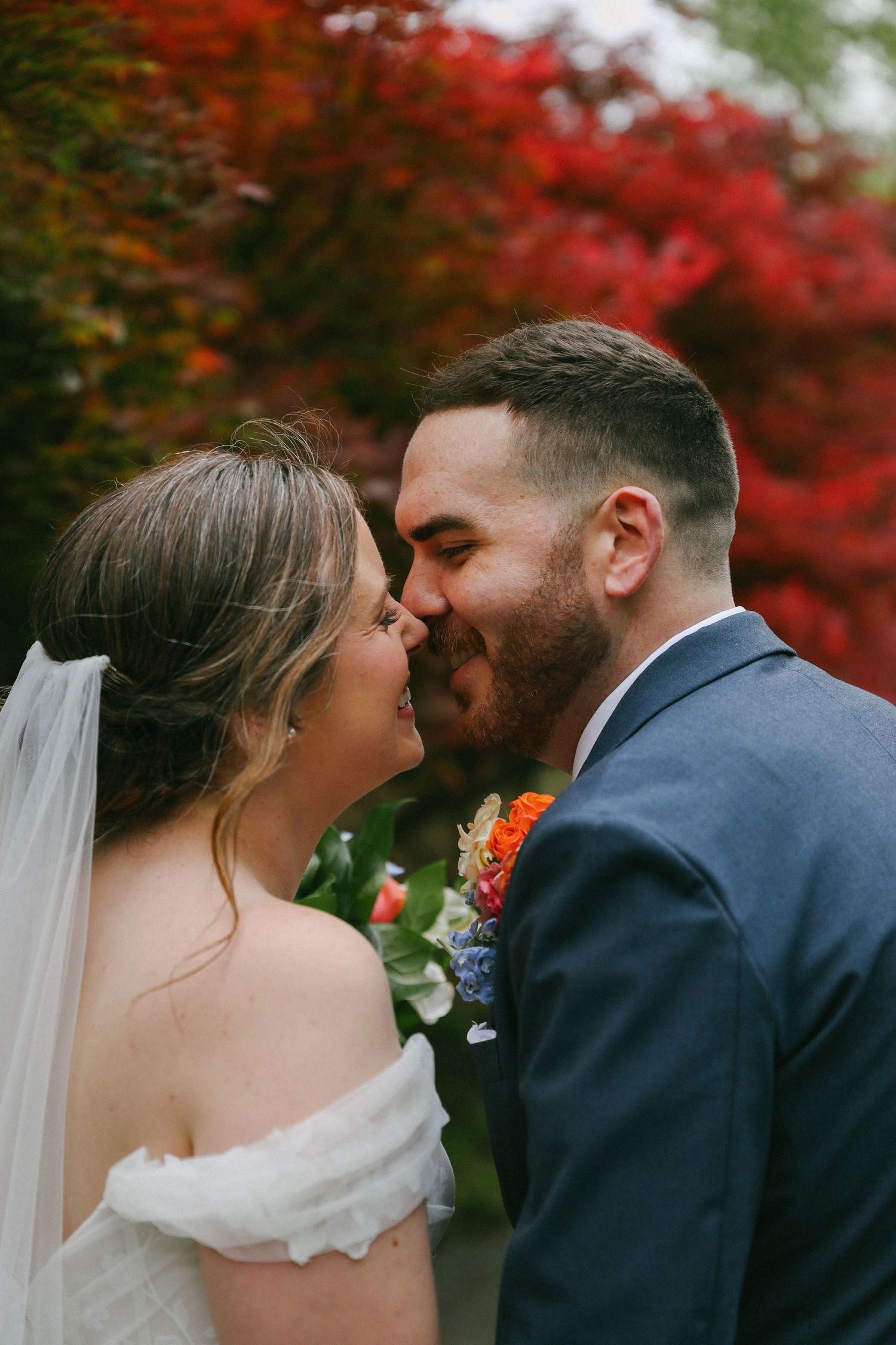Bearded groom in navy suit kissing bride in white dress with colorful boutonniere outdoors.