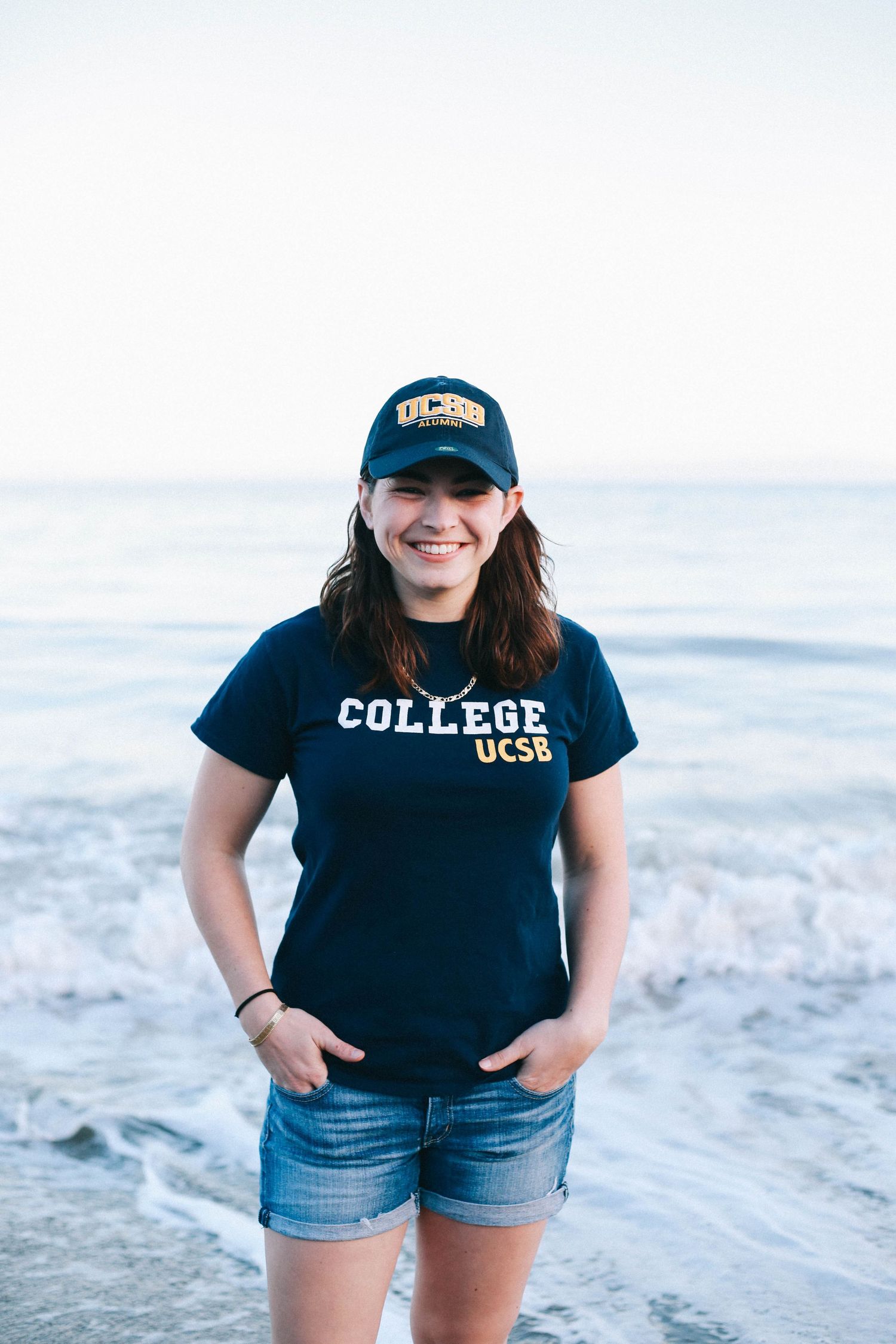 Happy woman in UCSB cap and t-shirt standing on a beach with ocean waves behind her.