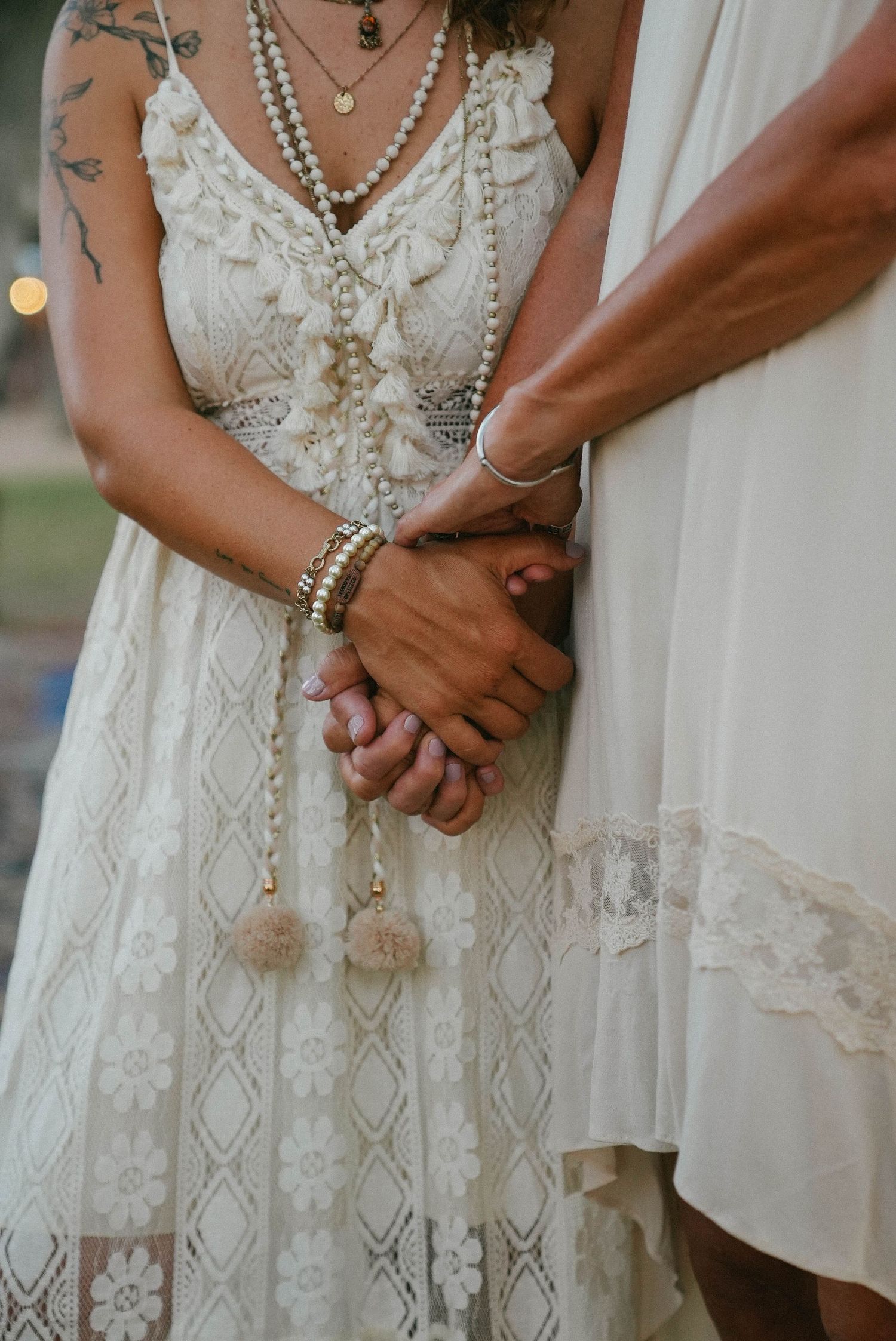 Close-up of two brides holding hands in boho lace wedding dresses with pearl jewelry.
