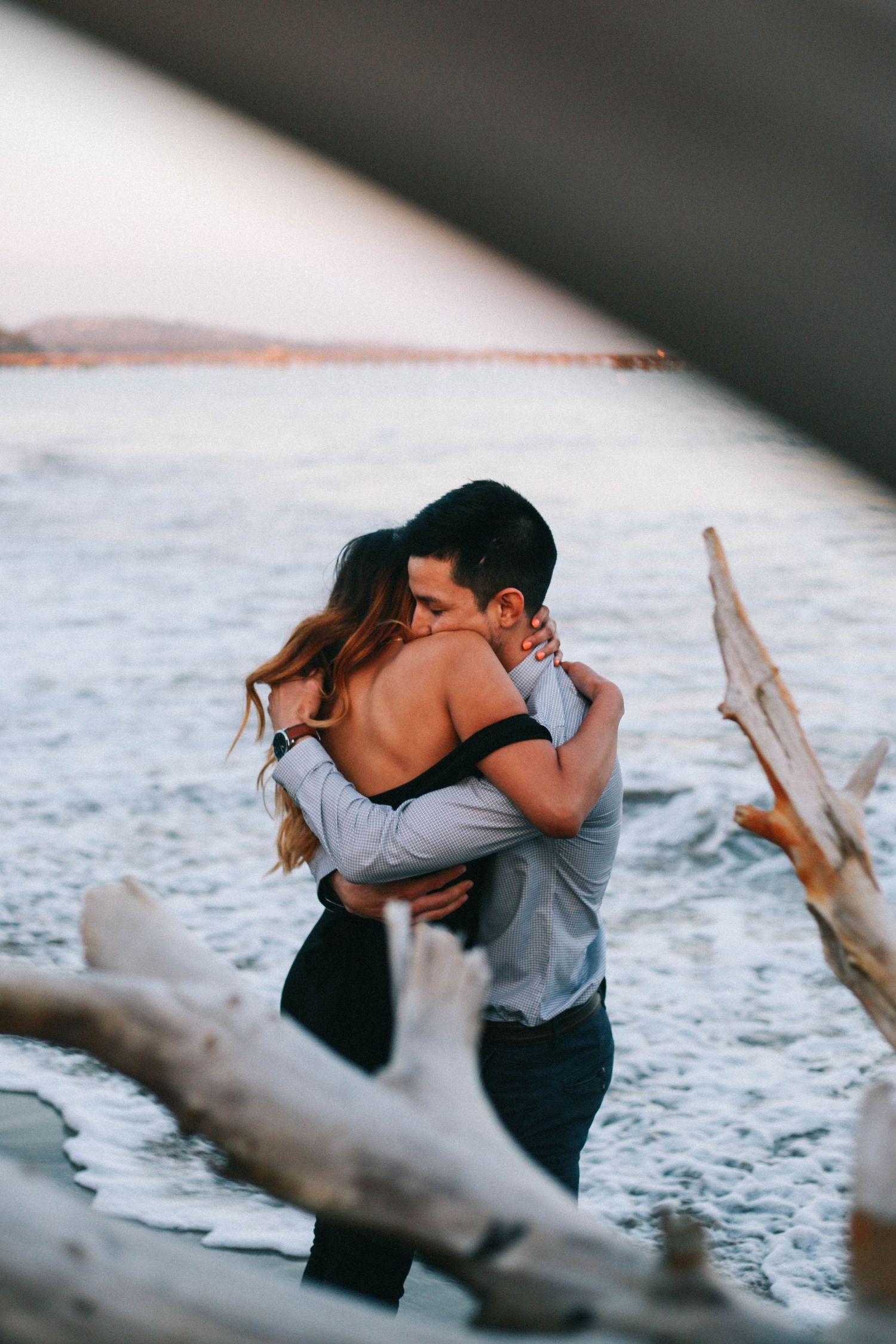 Romantic couple embracing and kissing near the water with driftwood in the foreground.
