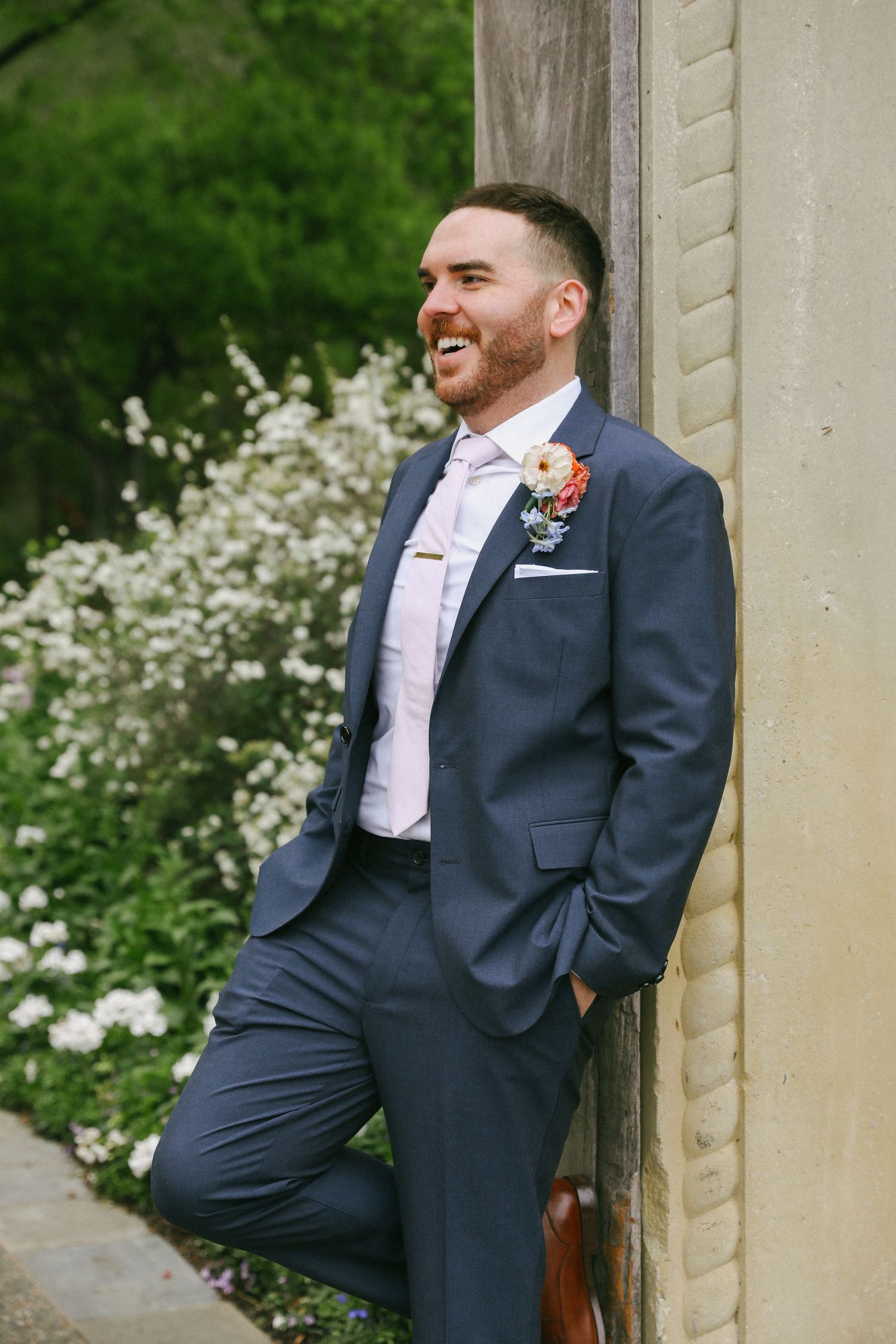 Smiling groom in navy suit with floral boutonniere leaning against stone pillar outdoors.