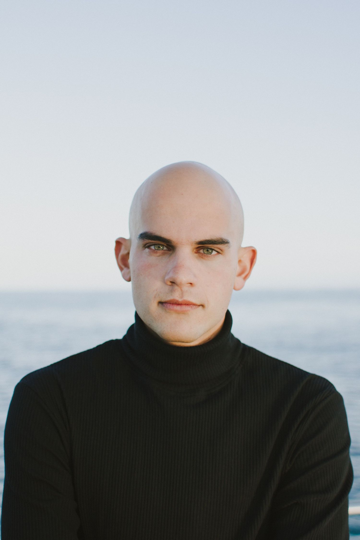 Bald young man in black turtleneck posing seriously with ocean horizon behind him.