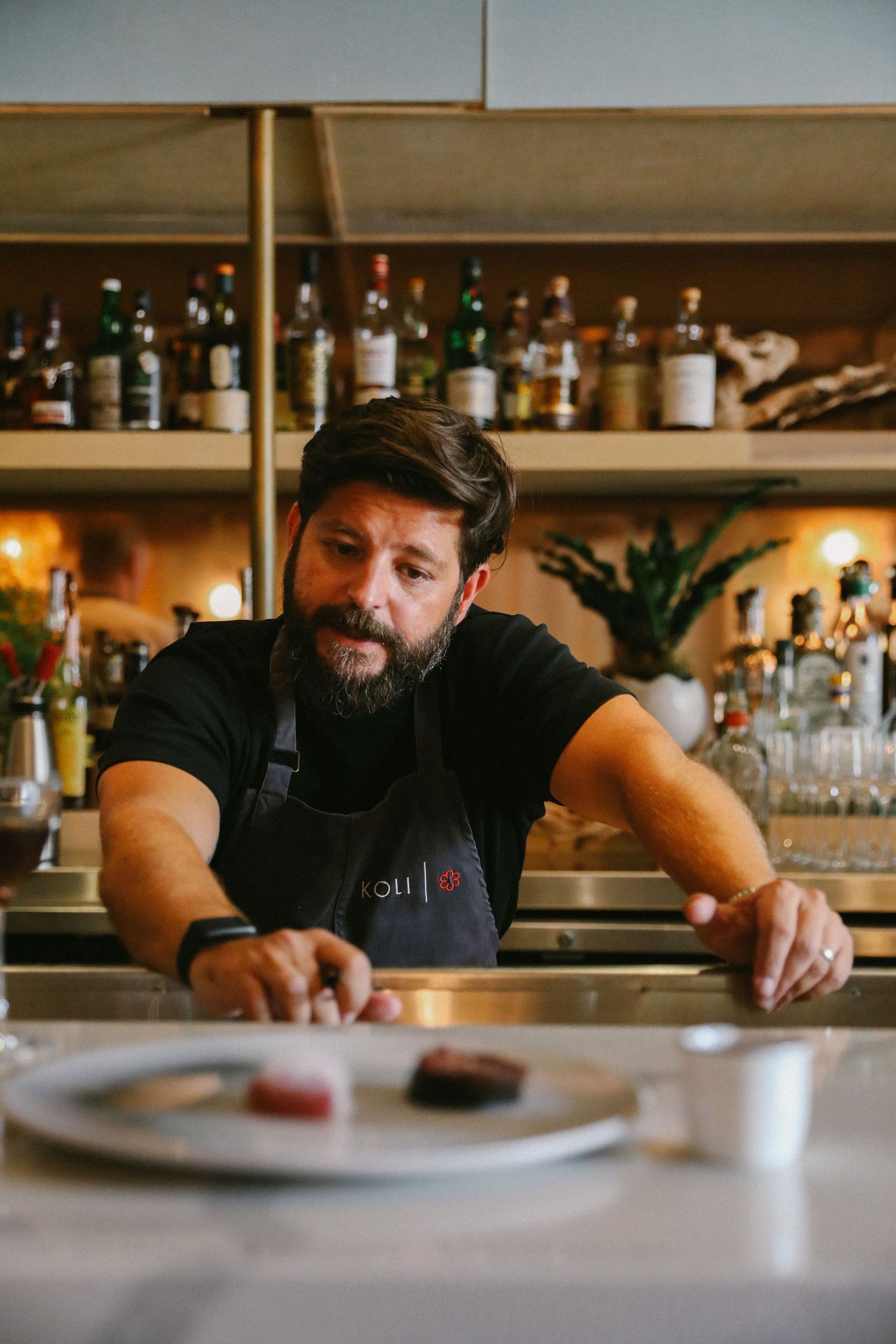 Bearded chef in black apron presenting plated dishes at modern restaurant bar counter.