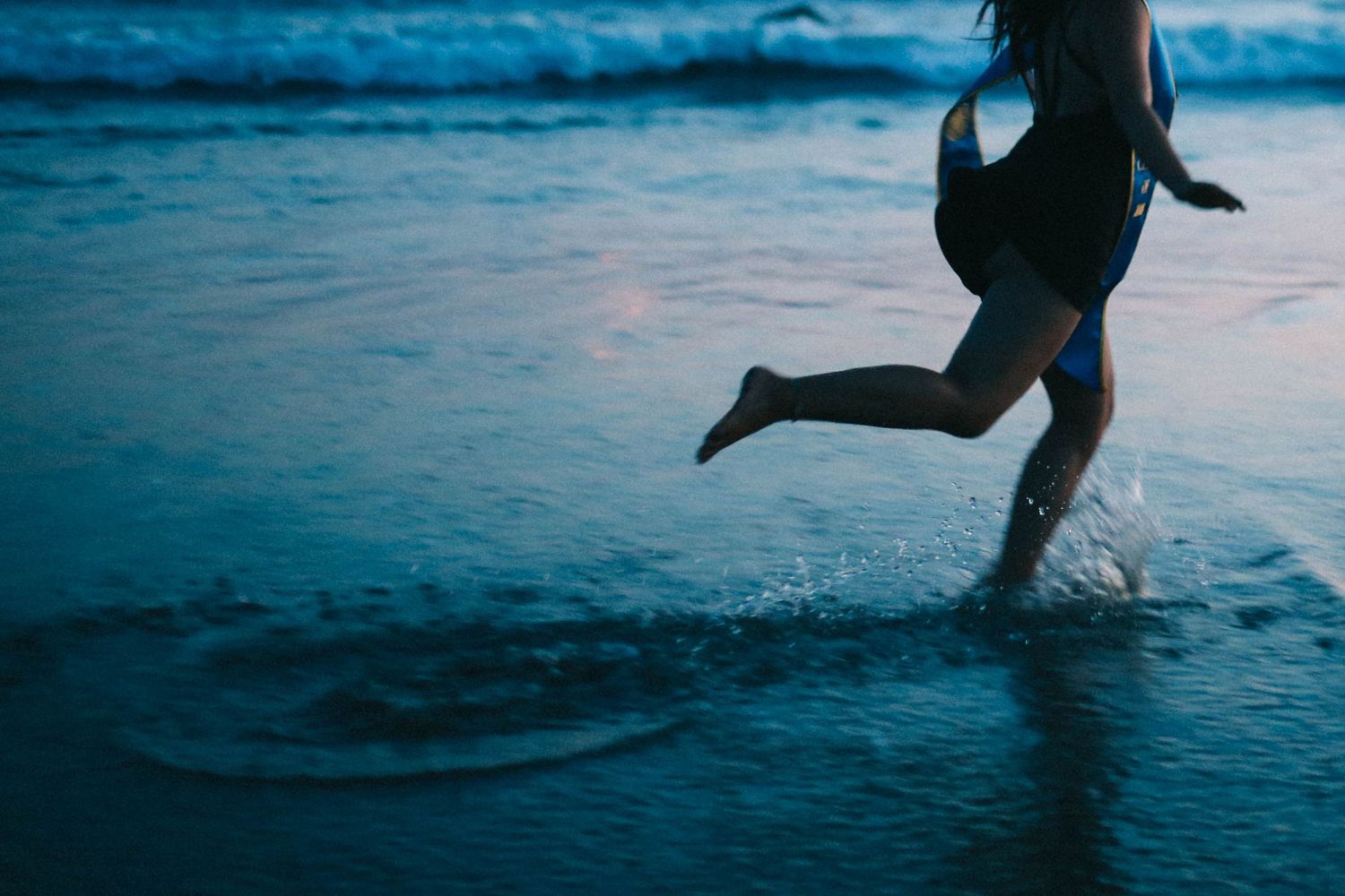 Silhouette of a person running barefoot through ocean waves at dusk on the beach.