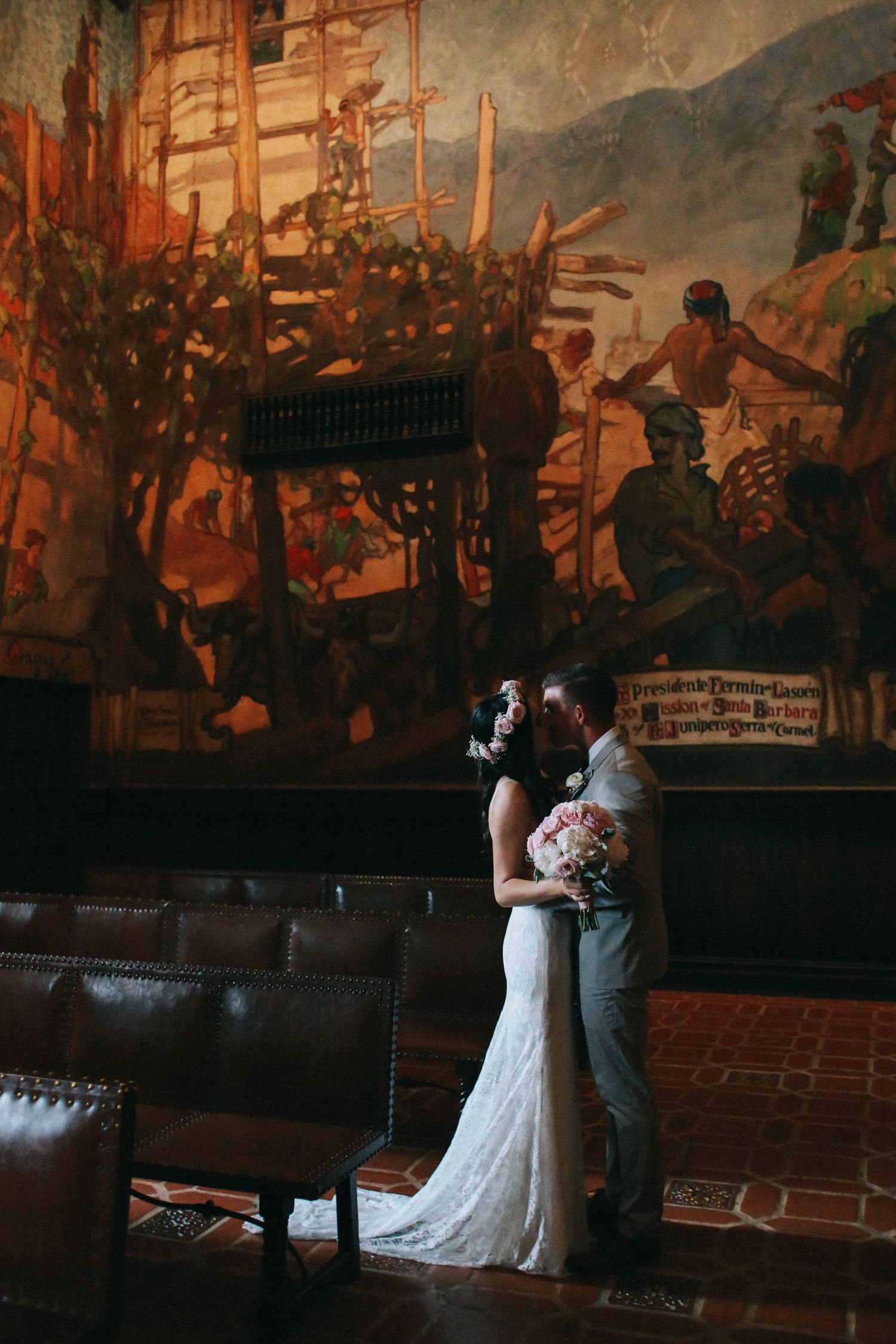 Bride and groom embracing in front of a large historic mural in an elegant wedding venue.