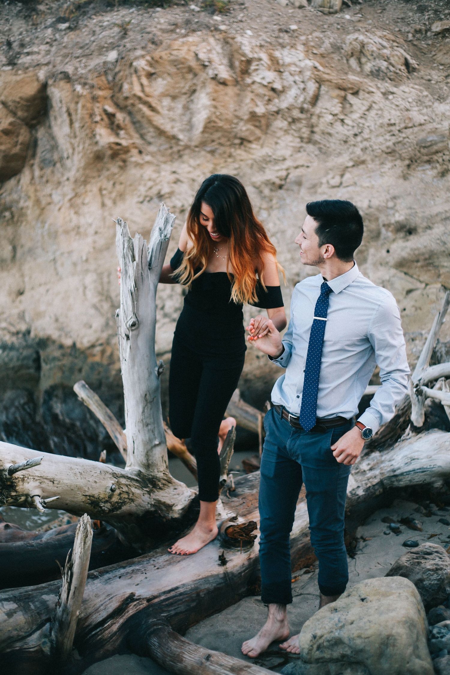 Couple holding hands on driftwood logs on rocky beach, woman in black dress, man in shirt and tie.