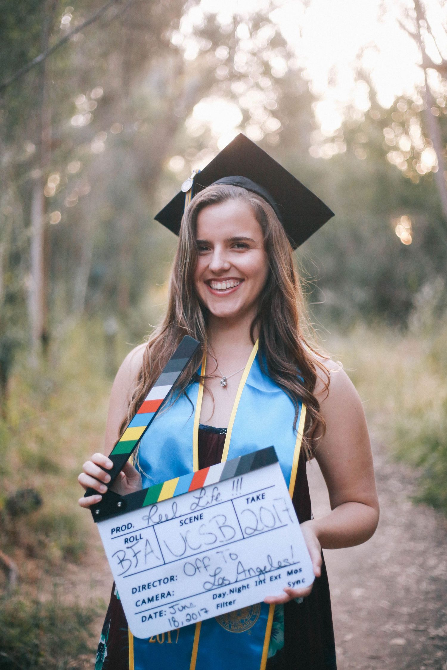 Smiling female graduate in cap and gown holding a film clapperboard outdoors.