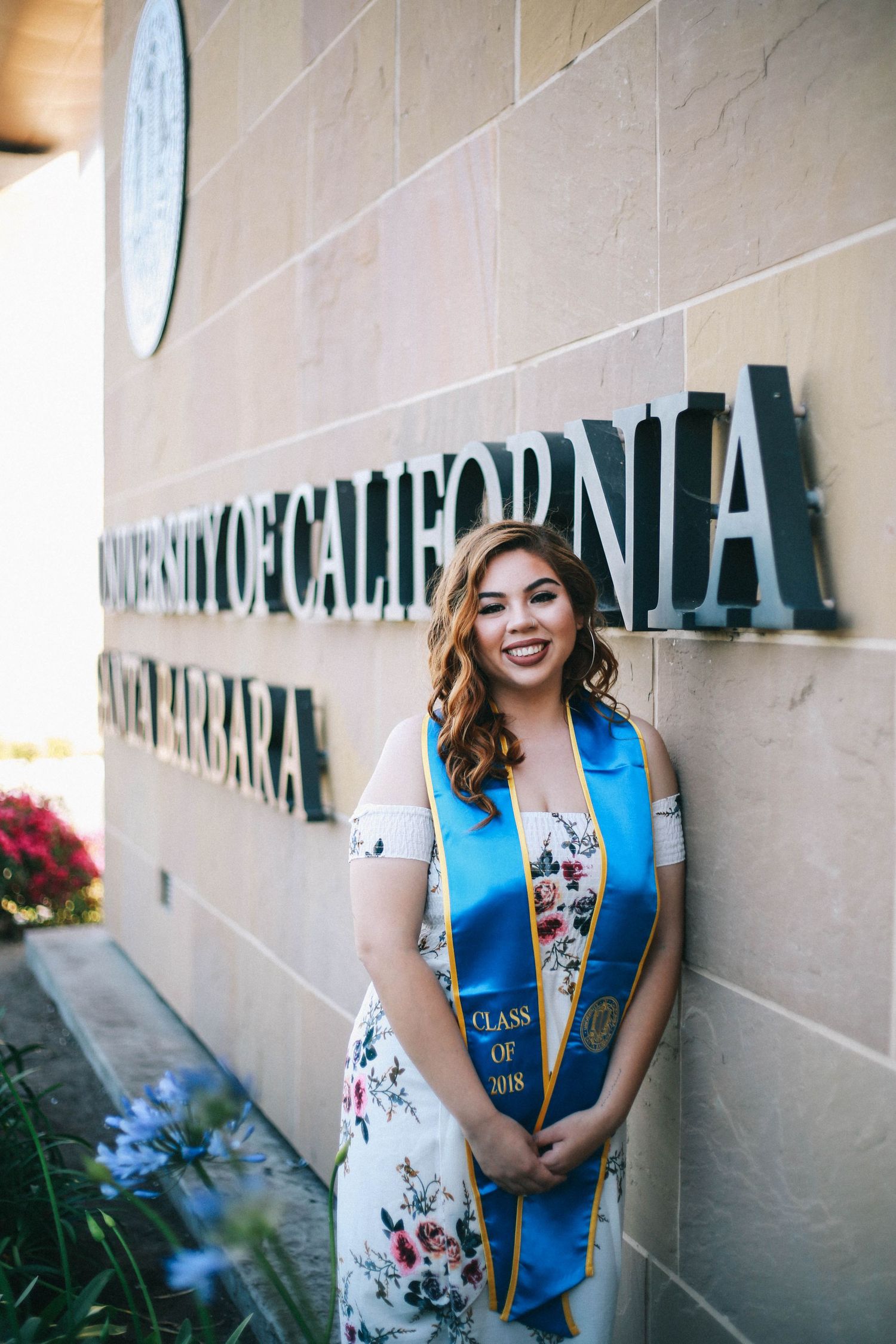 Smiling graduate in floral dress with UCSB Class of 2018 blue stole at campus wall.