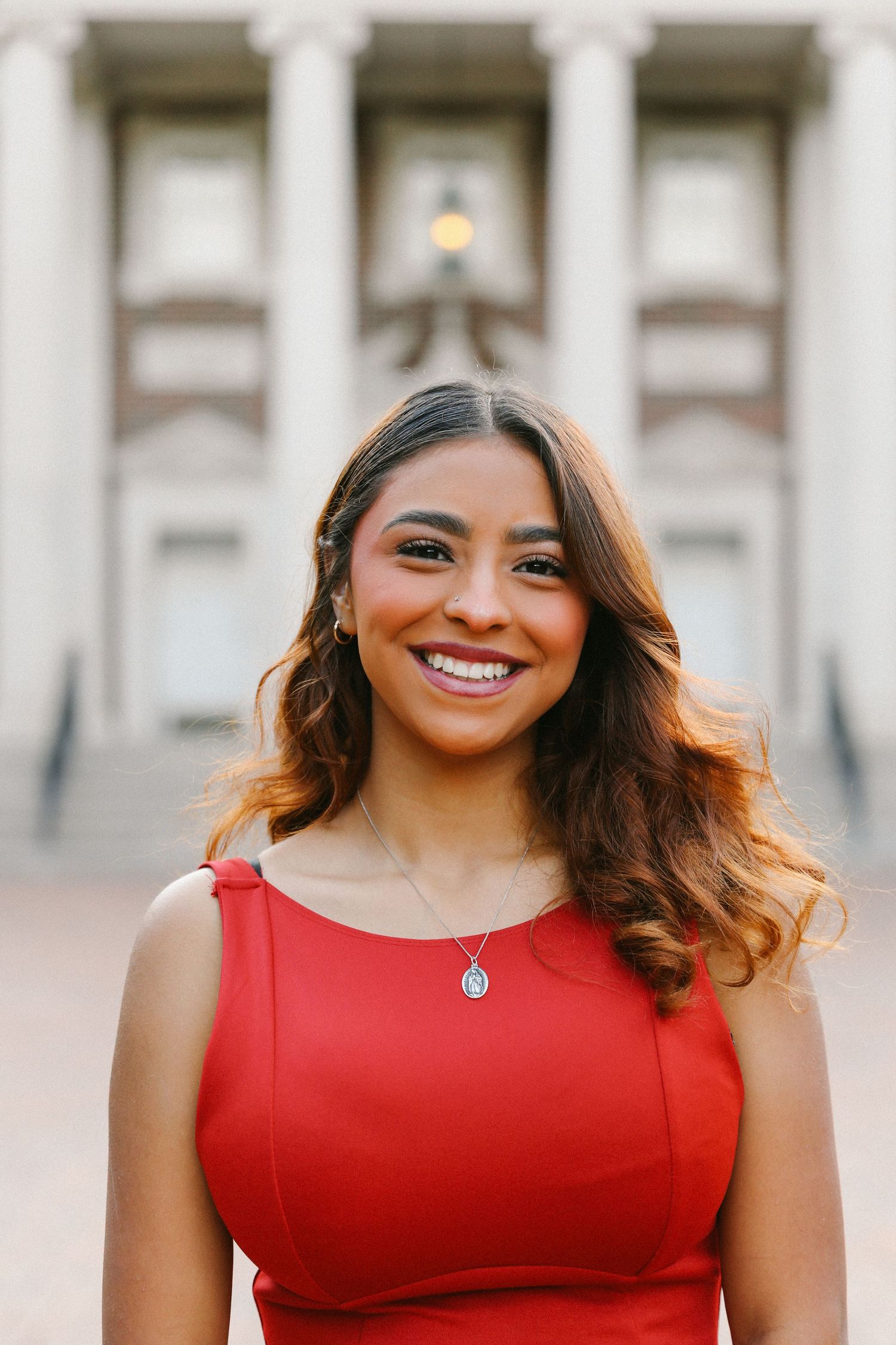 Smiling young woman in red dress with necklace posing in front of a columned building.