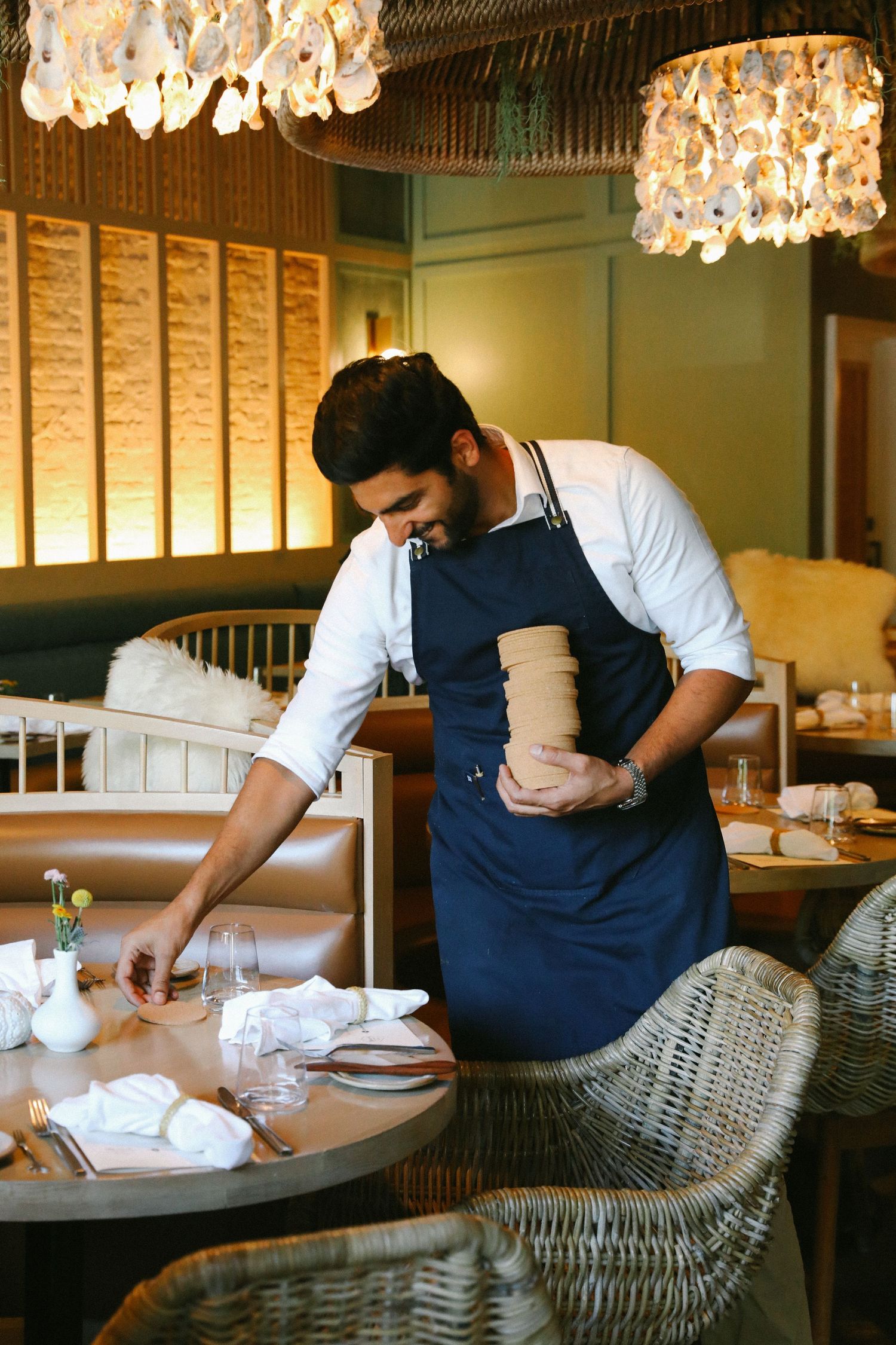 Restaurant staff member in blue apron setting table in warmly lit upscale dining room.