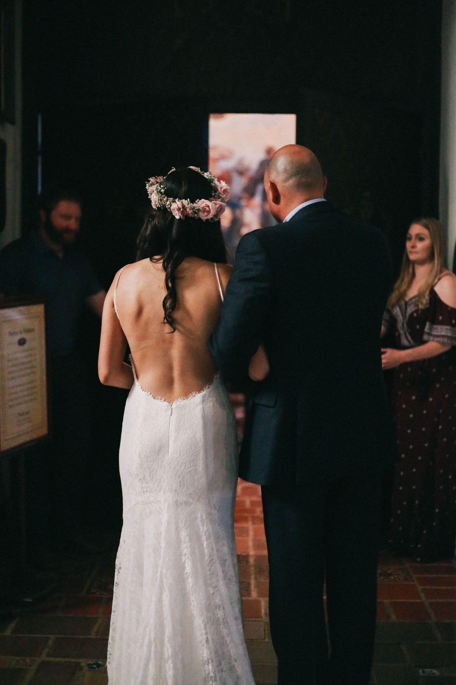 Bride in backless lace wedding gown with floral crown standing with groom during ceremony.