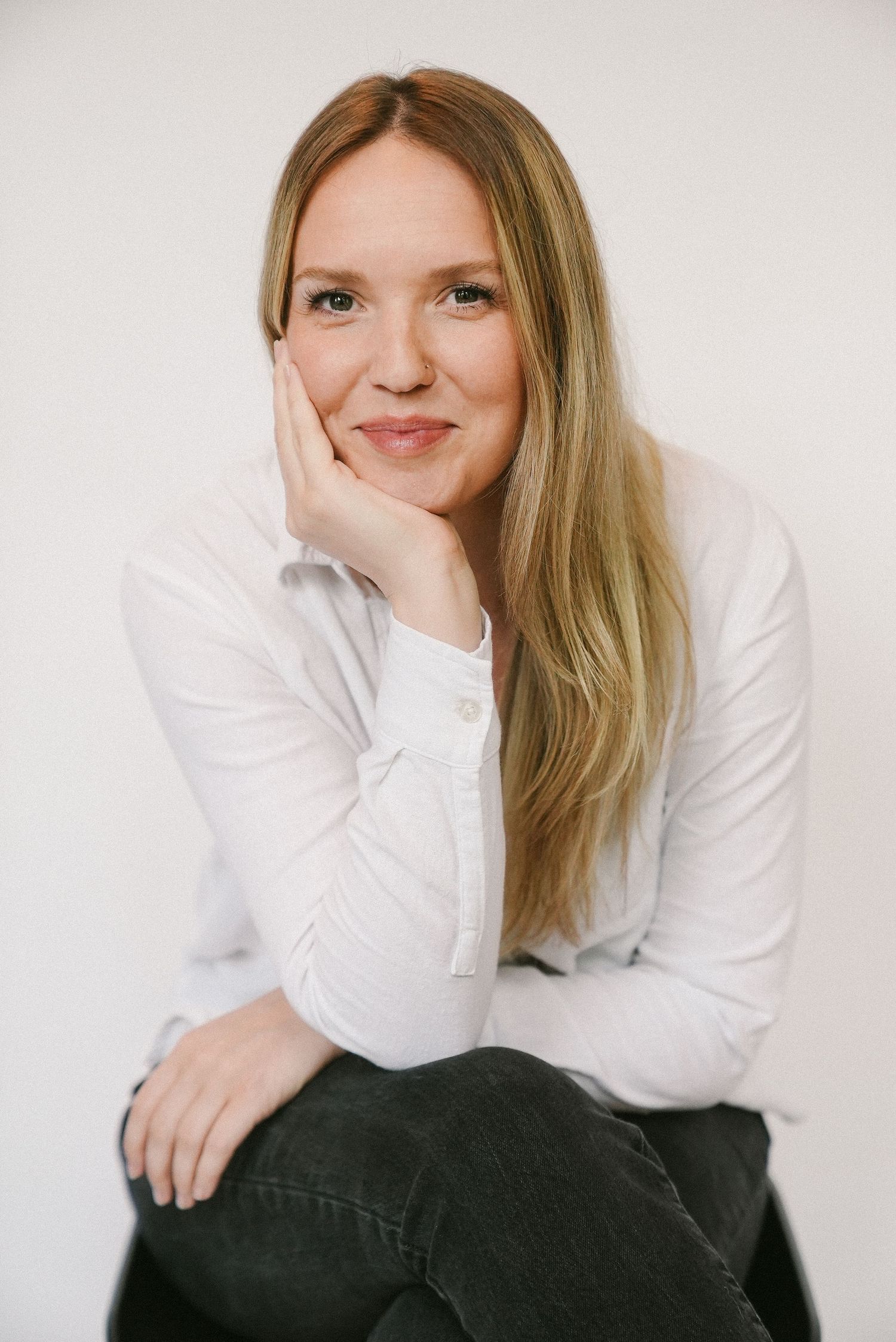 Smiling blonde woman in white shirt sitting relaxed against a white studio background.