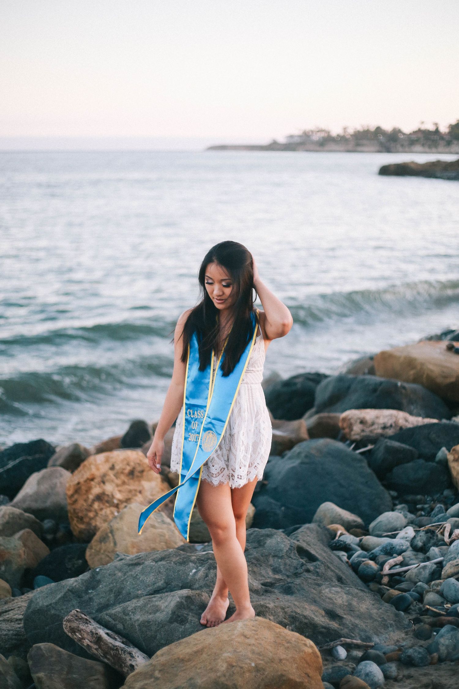 Young woman in white dress with UCSB blue graduation stole standing on rocky shoreline.