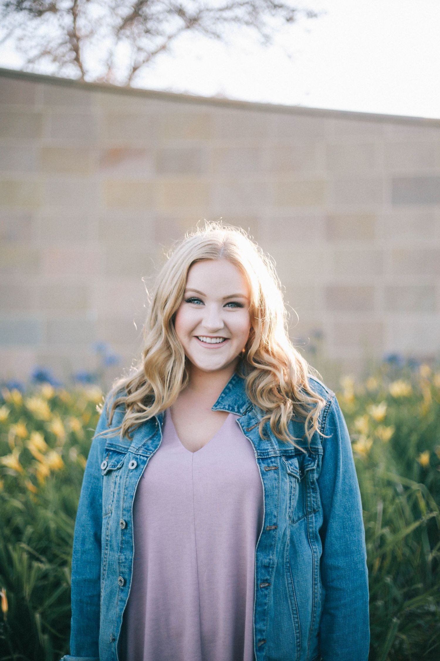 Smiling blonde woman in denim jacket and pink dress standing in front of a stone wall with flowers.