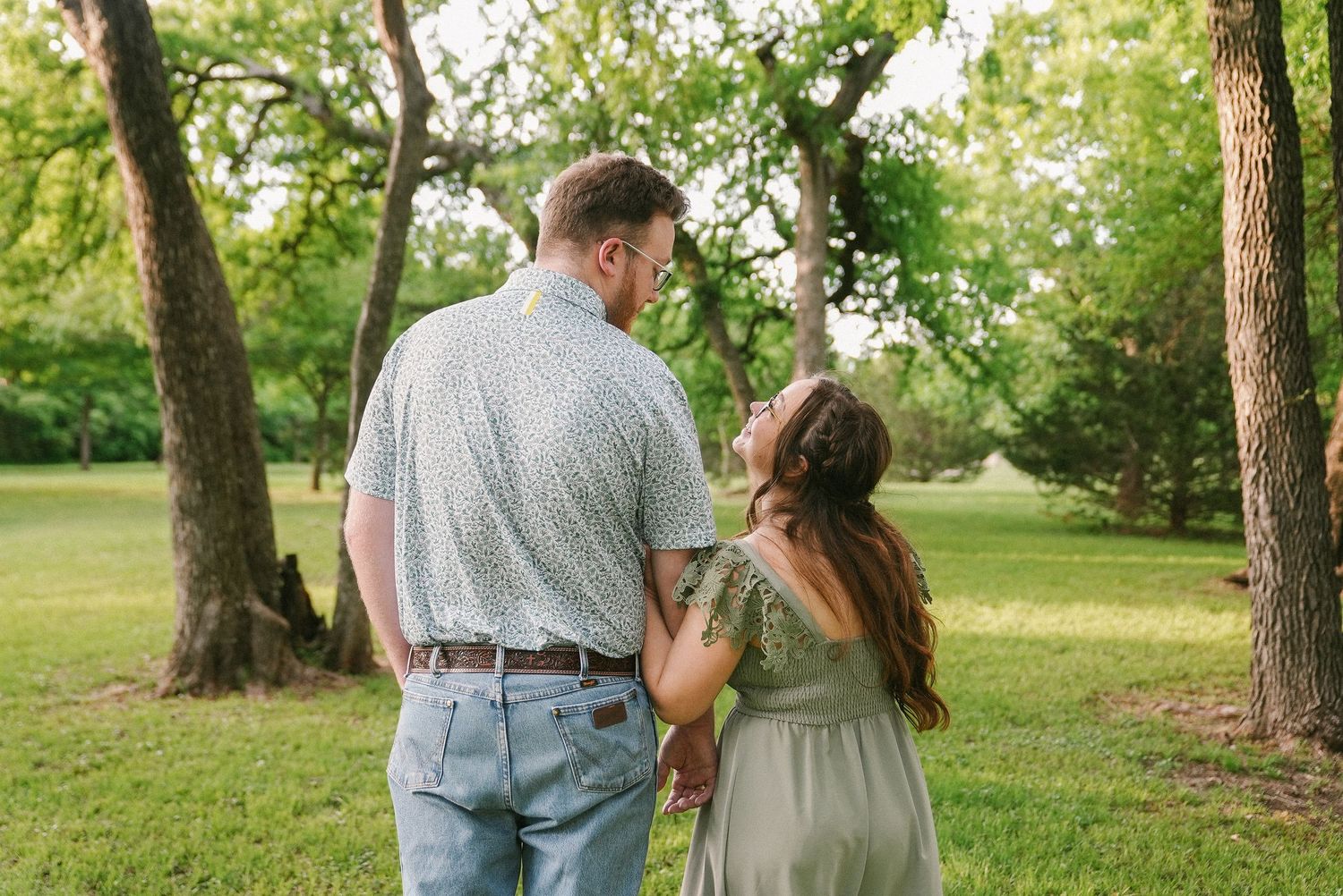 Couple holding hands and gazing at each other in a lush green park setting.