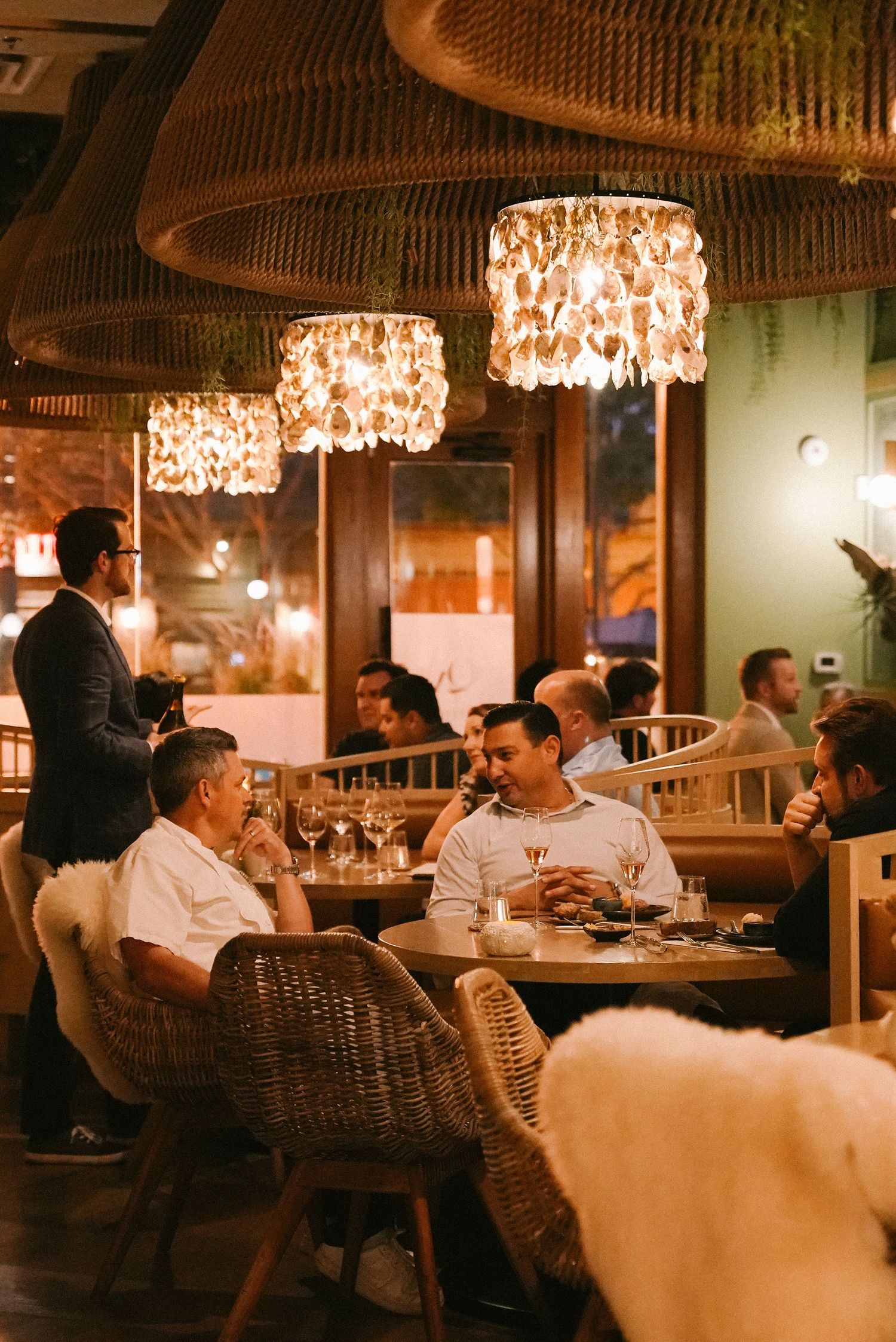 Lively fine dining restaurant filled with guests enjoying meals beneath decorative rattan chandeliers.