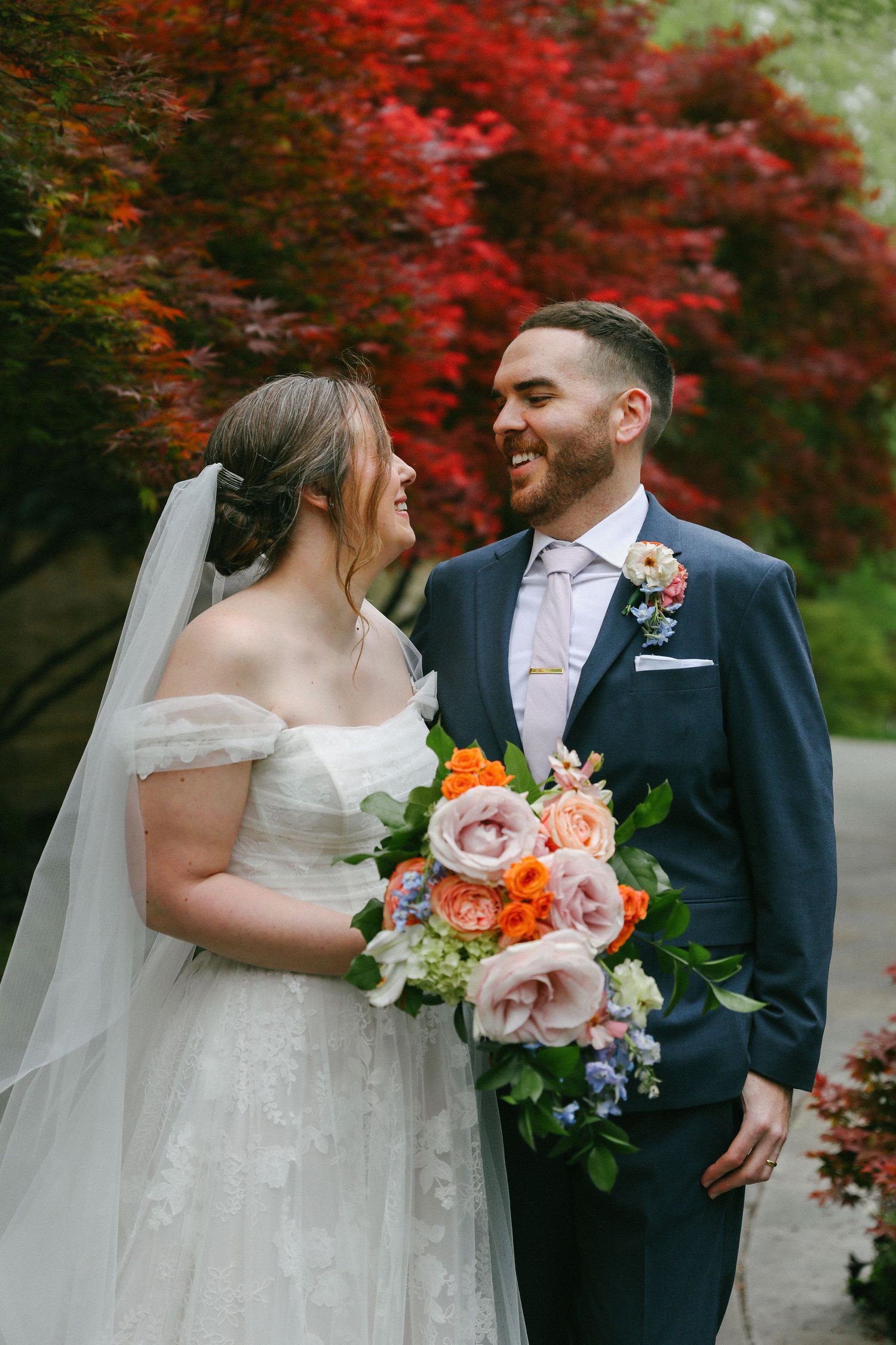 Bride and groom smile at each other in a garden with vibrant red Japanese maple trees in the background.