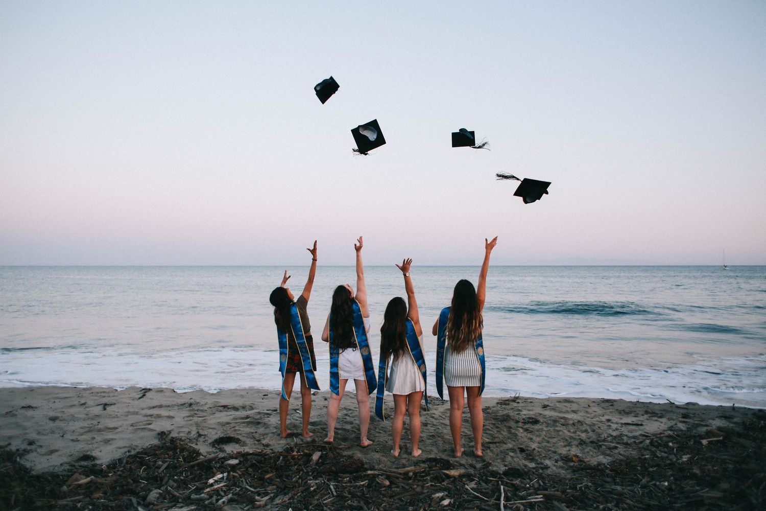 Four graduates toss caps in the air on a beach at dusk celebrating their achievement.
