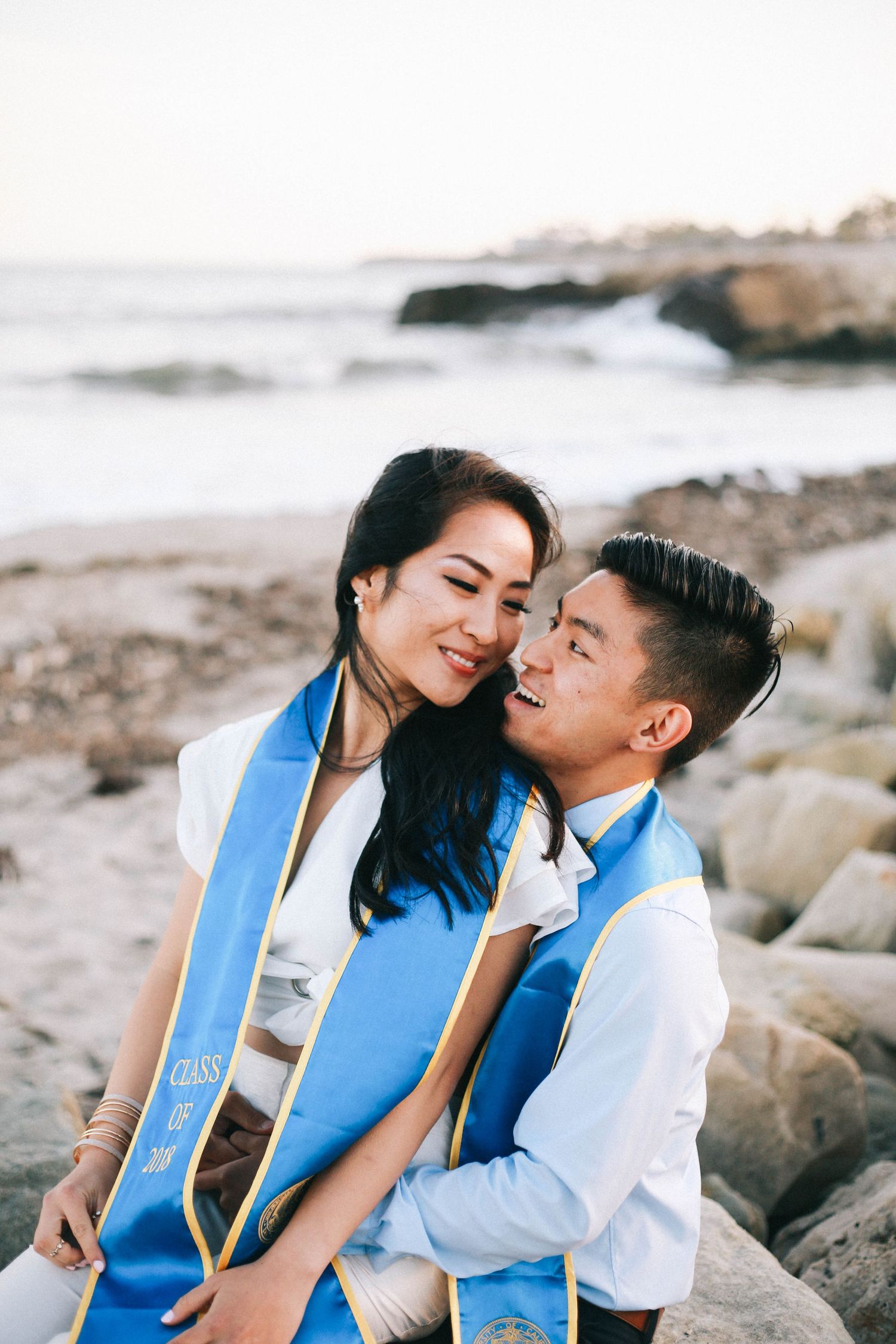 Two graduates in blue and gold stoles sit together smiling on rocky beach at sunset.