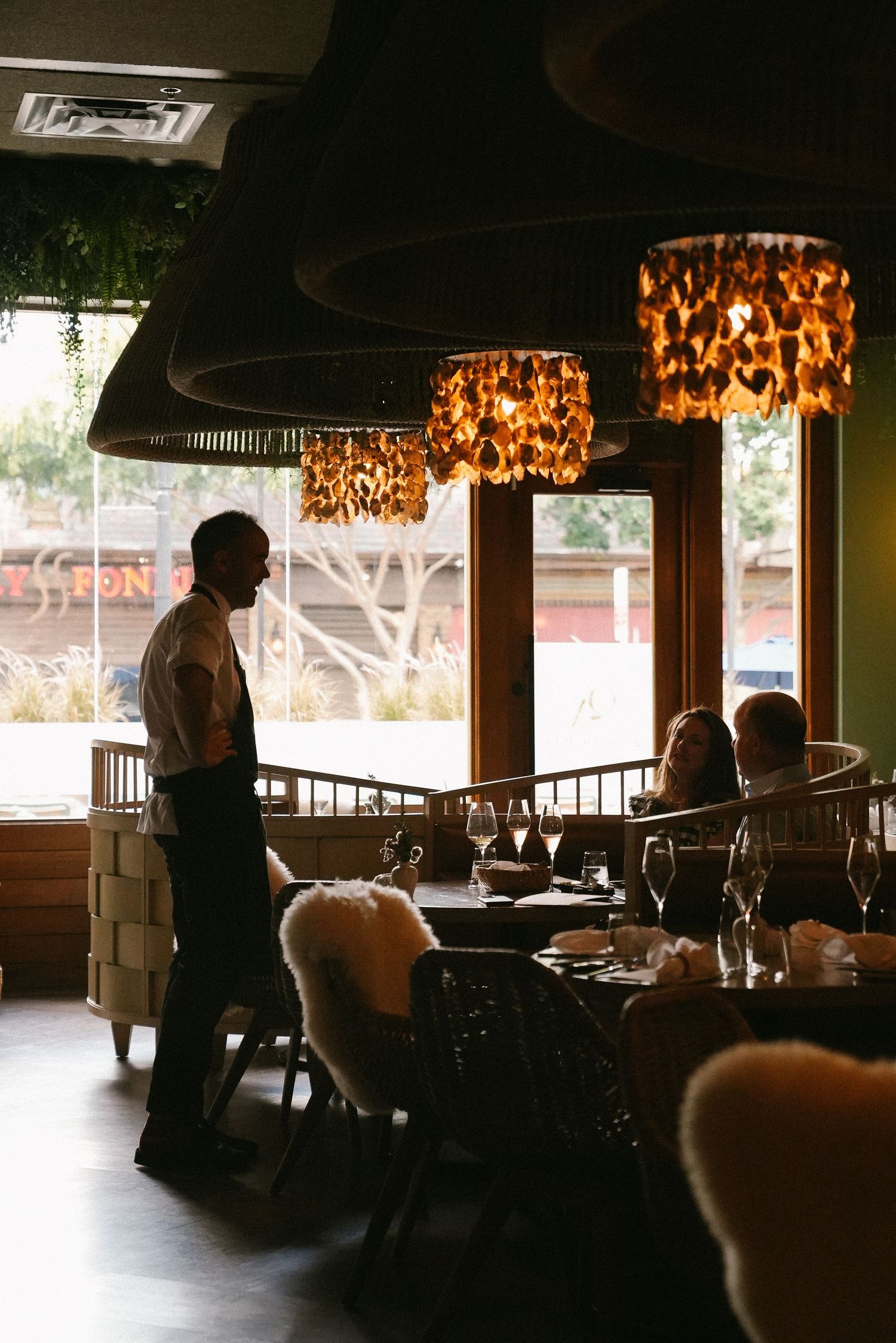 Waiter serves guests at candlelit tables beneath ornate amber pendant lights in upscale restaurant.