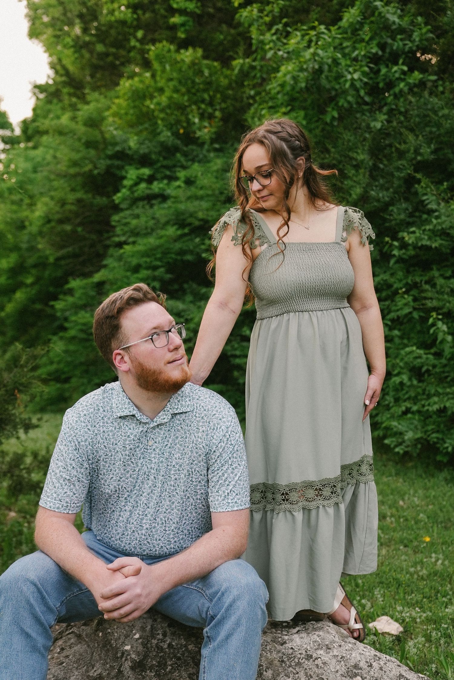 Couple poses on rocks outdoors, man sitting while woman in sage green dress stands beside him.