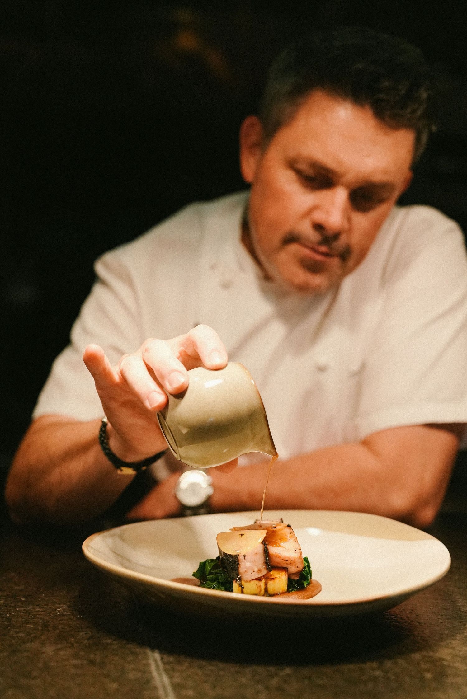 Chef in white jacket carefully pours sauce over a plated gourmet dish in a fine dining kitchen.
