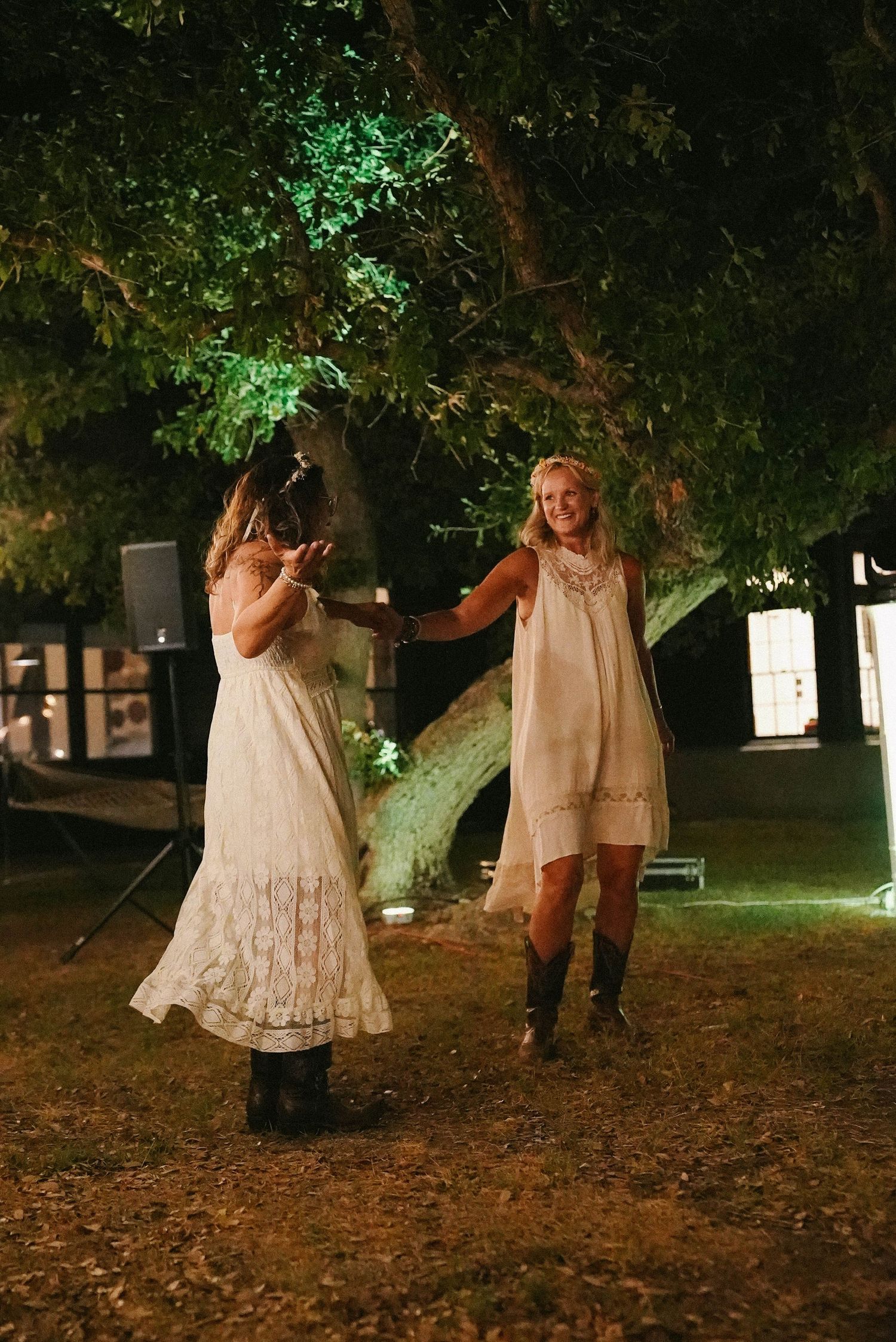 Two brides in white dresses and cowboy boots dance together under illuminated trees at a night wedding.