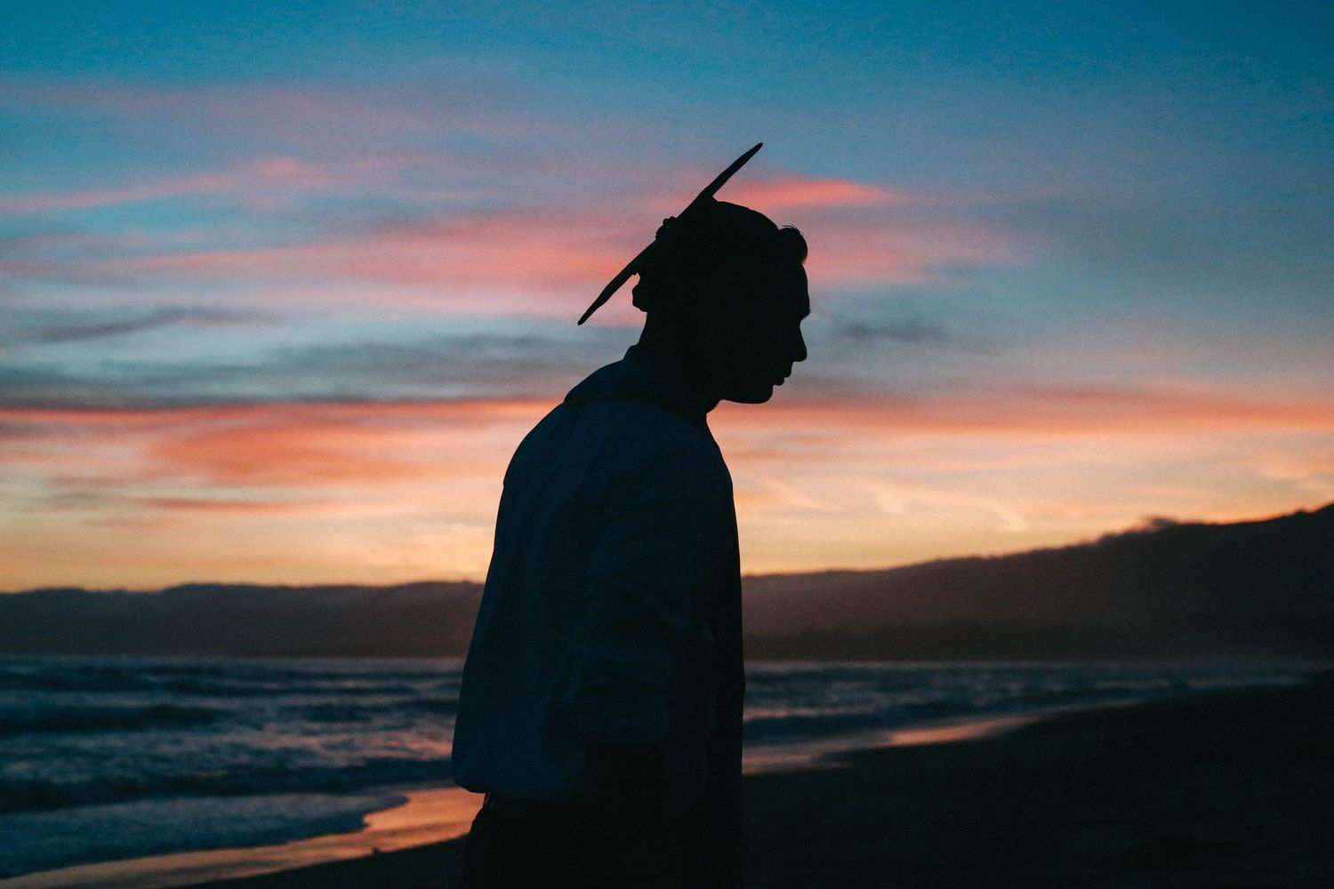 Silhouette of a person with feathers in hair standing on a beach at colorful sunset.