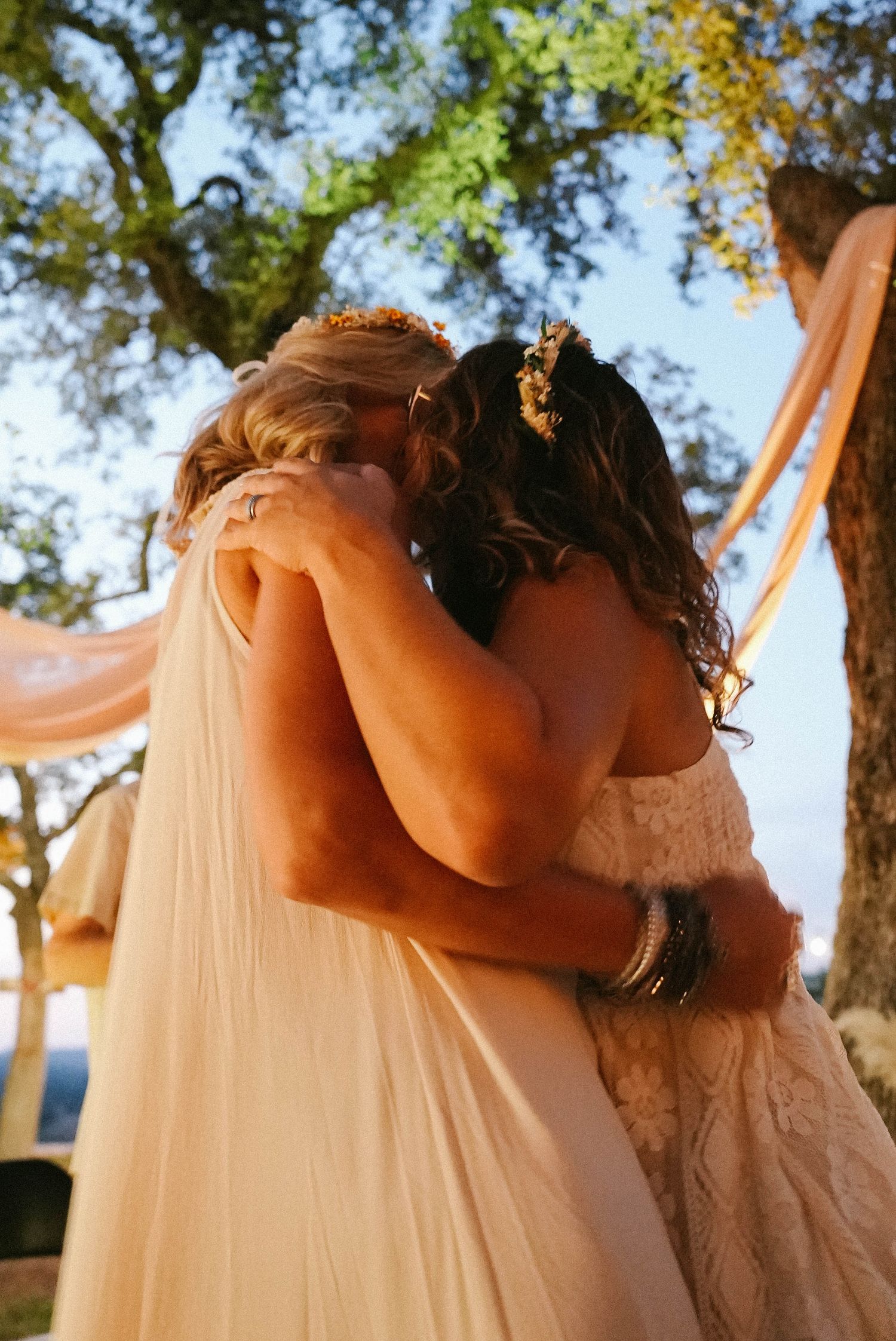 Two brides sharing emotional first kiss under draped fabric at outdoor bohemian wedding.