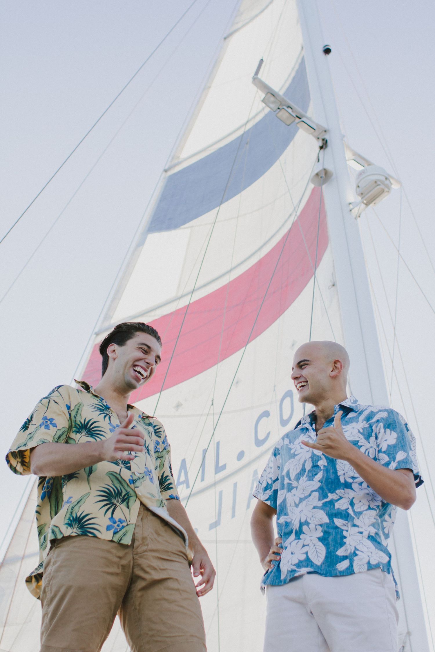 Two smiling men in floral shirts standing in front of a sailboat sail outdoors.