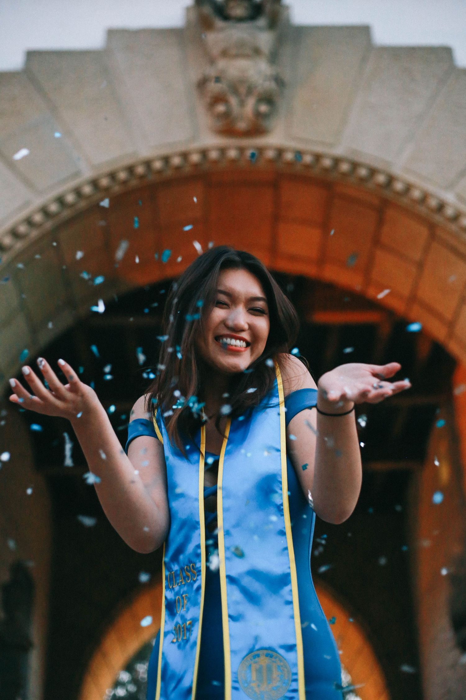 Smiling woman in blue graduation regalia celebrating with blue confetti under an archway.