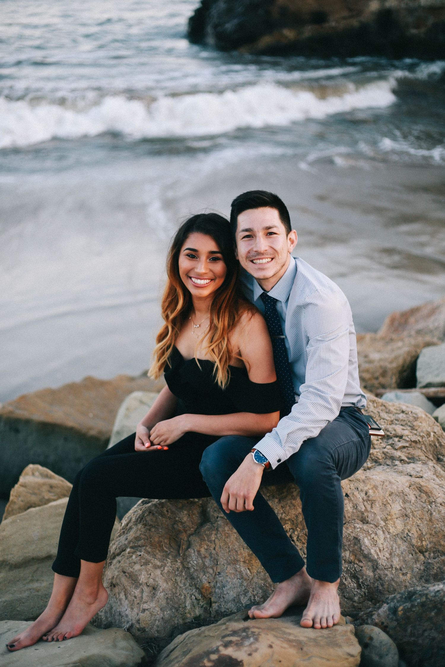 Smiling couple sitting on rocks at the beach, woman in black dress and man in gray shirt and tie.