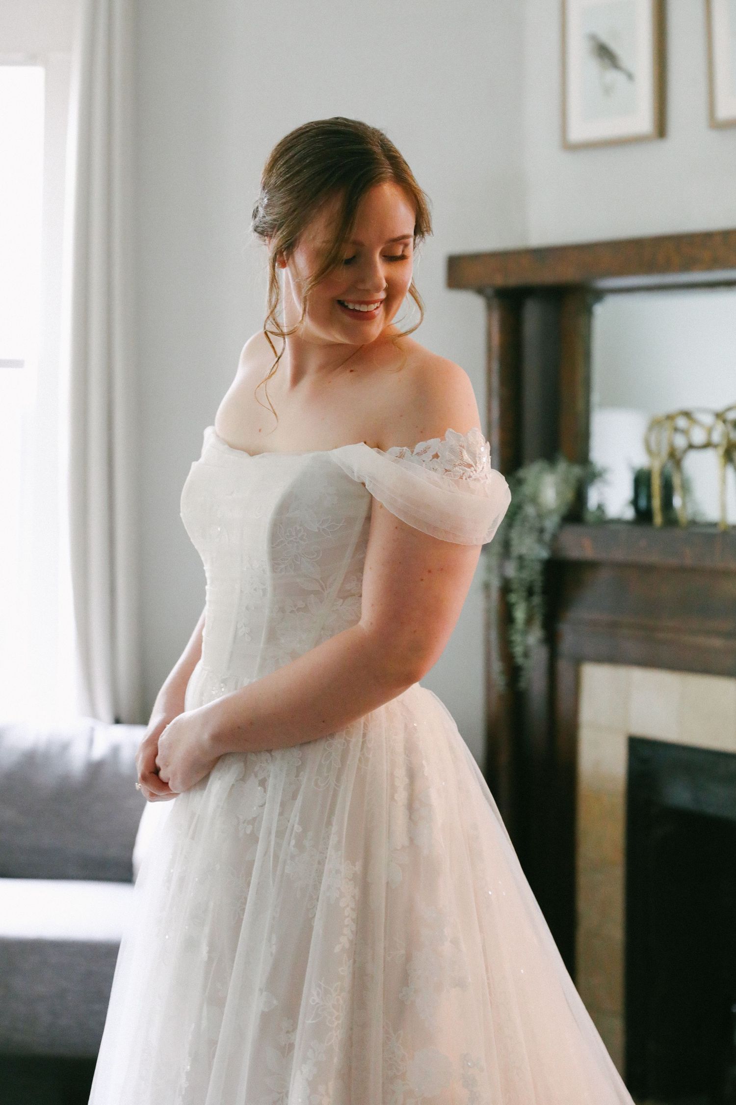Bride smiles in an off-shoulder lace ball gown while getting ready in a beautifully decorated room.