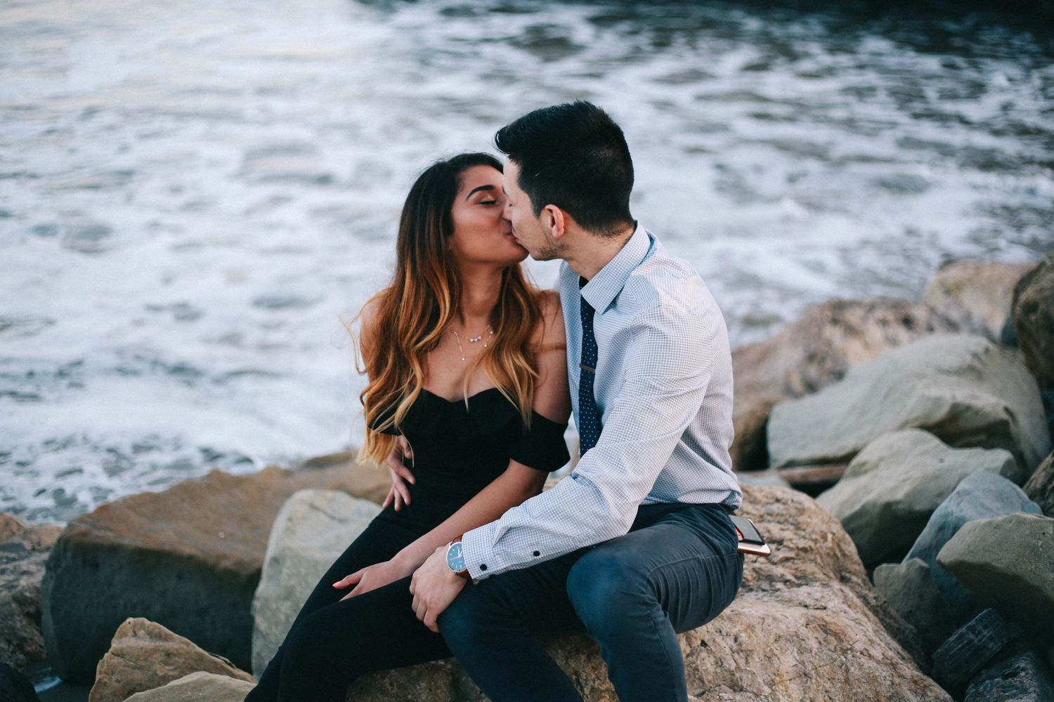 A couple shares a romantic kiss while sitting on rocky seaside boulders with ocean waves behind them.