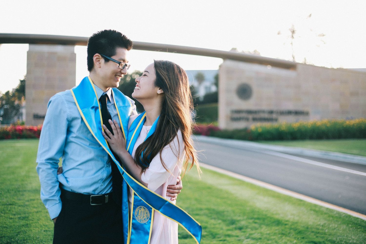 Couple in graduation stoles embracing and smiling at each other on a university campus lawn.