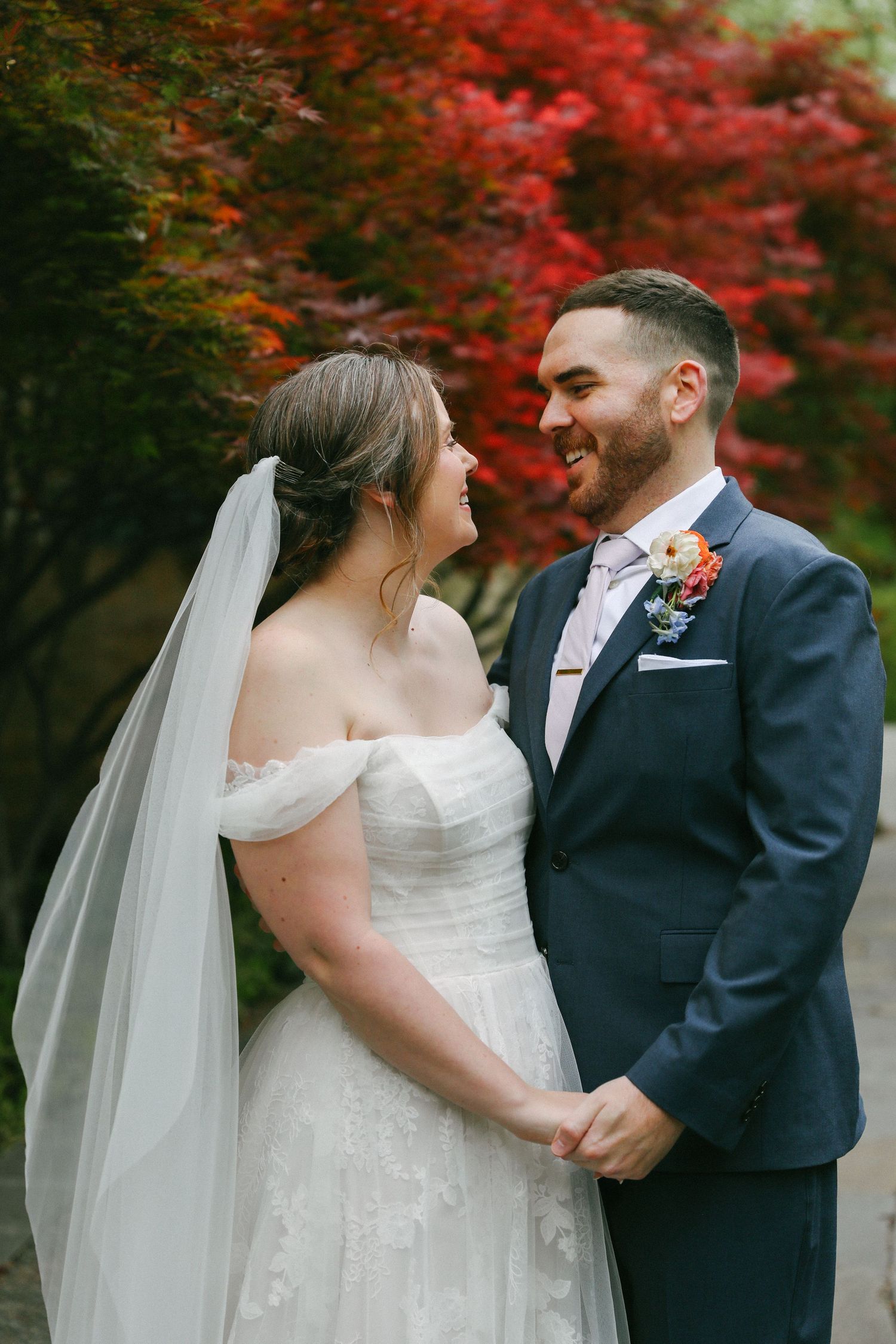 Bride in off-shoulder lace gown and groom in navy suit smile warmly amid vibrant red autumn foliage.