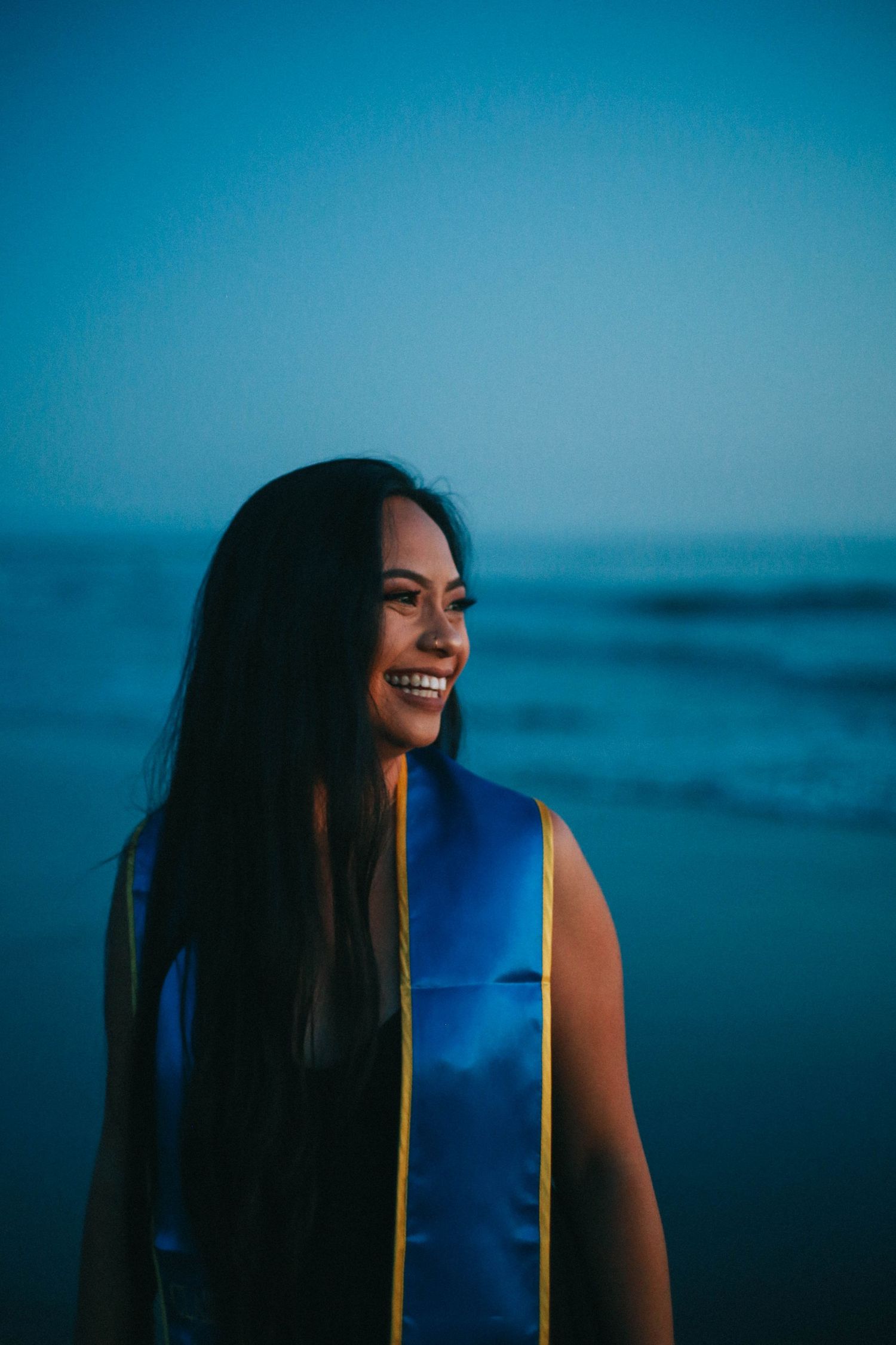Smiling young woman wearing a blue UCSB graduation stole standing by the ocean at twilight.