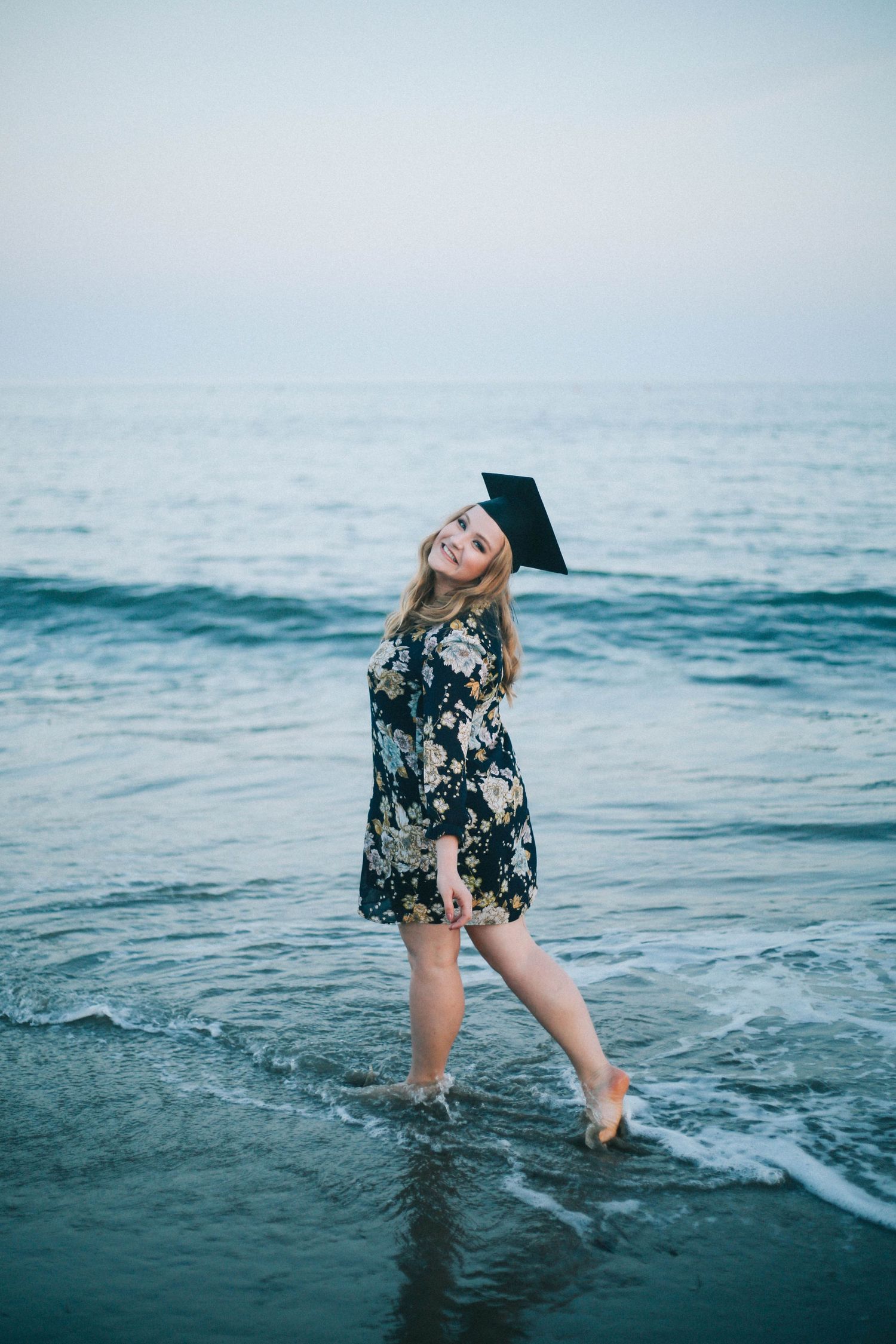 Smiling blonde woman in floral dress and graduation cap wading in ocean waves at the beach.