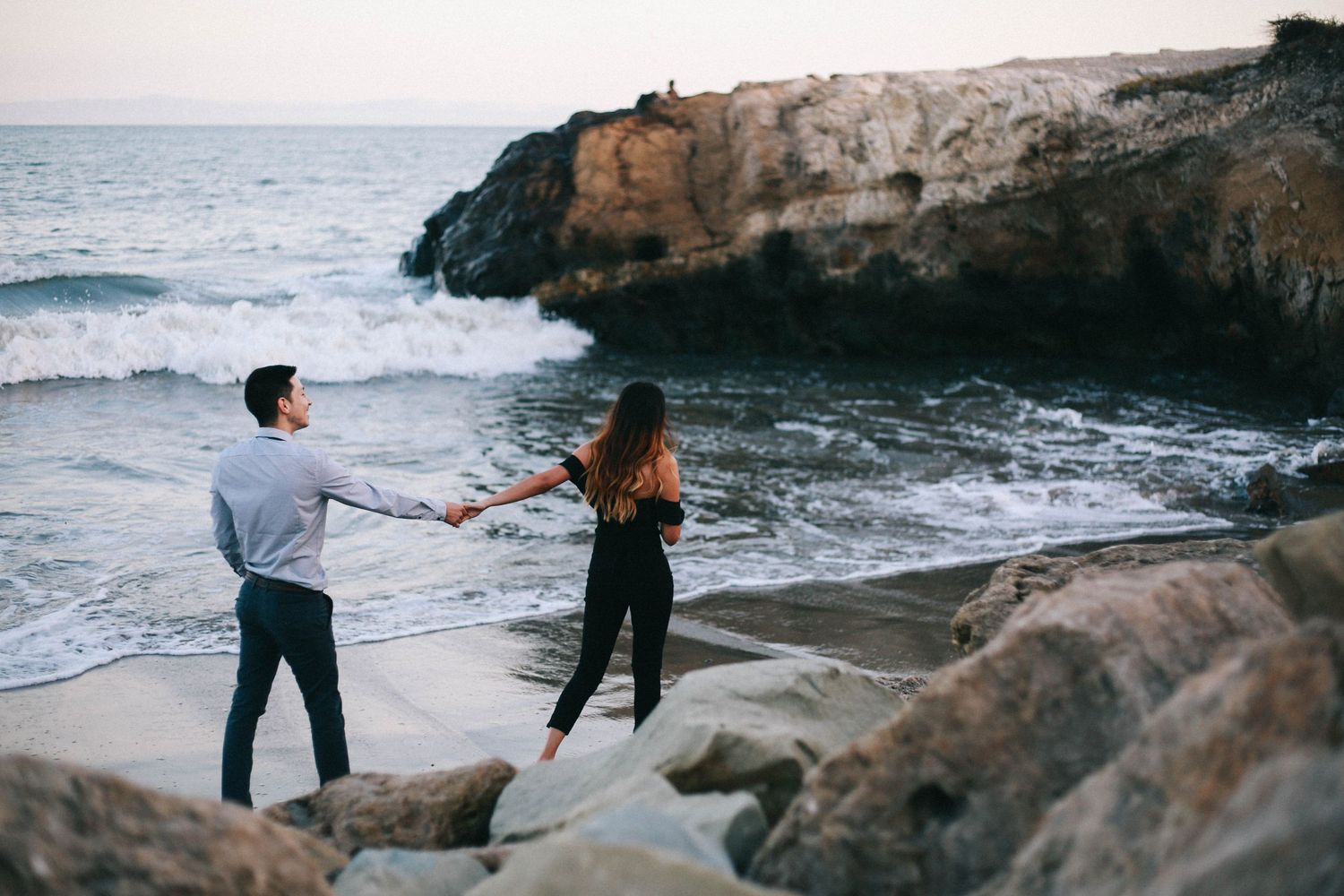 Couple holds hands on rocky coastal cliffs with dramatic ocean waves crashing in the background.