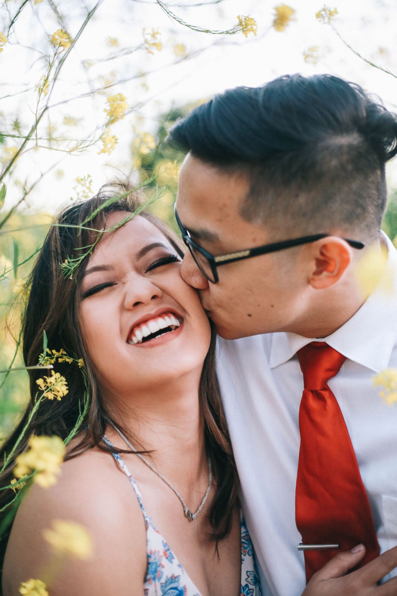 A young man kisses his laughing girlfriend on the cheek while surrounded by yellow wildflowers outdoors.